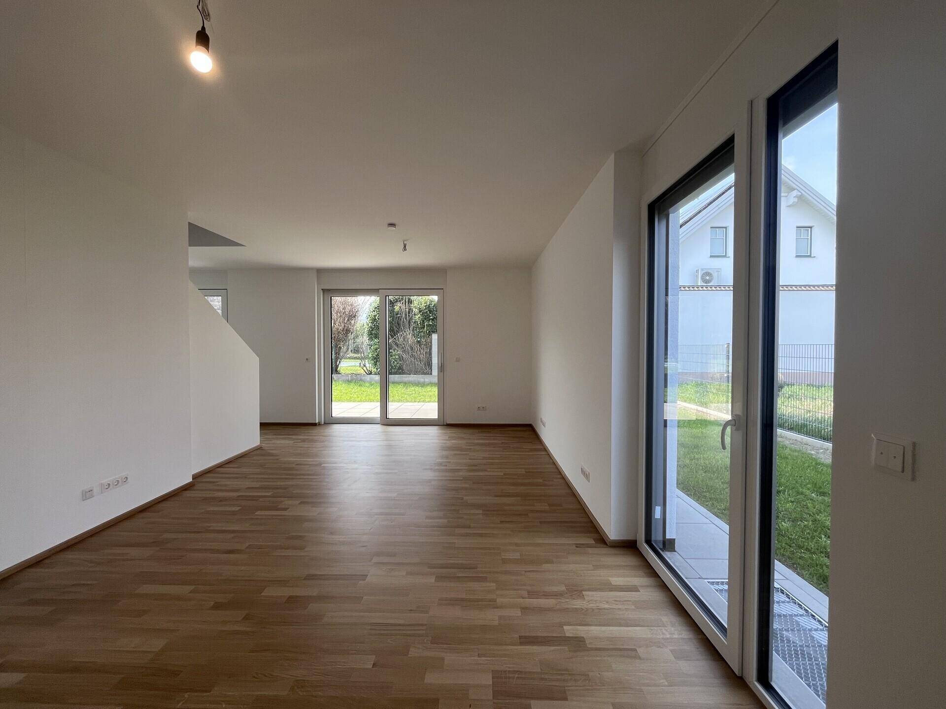 Geräumiges Wohnzimmer mit hellem Parkett und viel Tageslicht durch große Fenster.