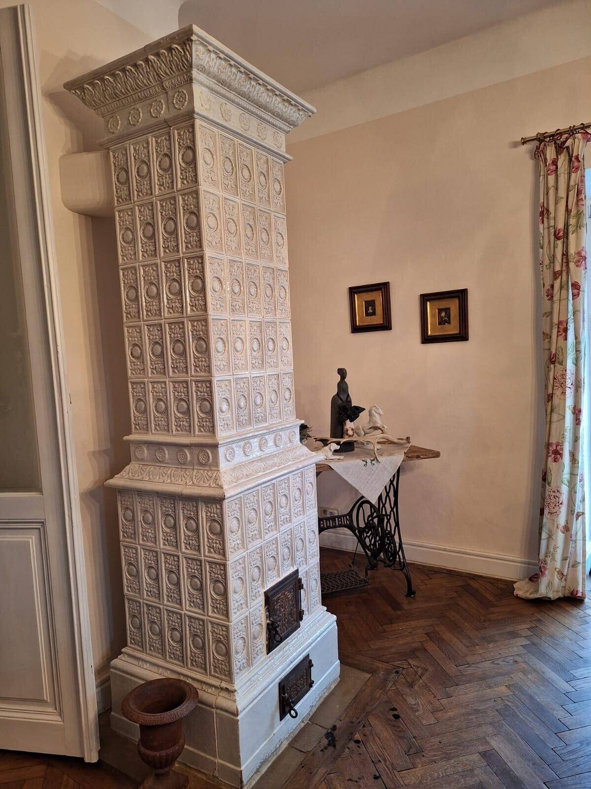 Hoher, weißer Kachelofen mit kunstvollen Verzierungen in einem traditionellen Raum.