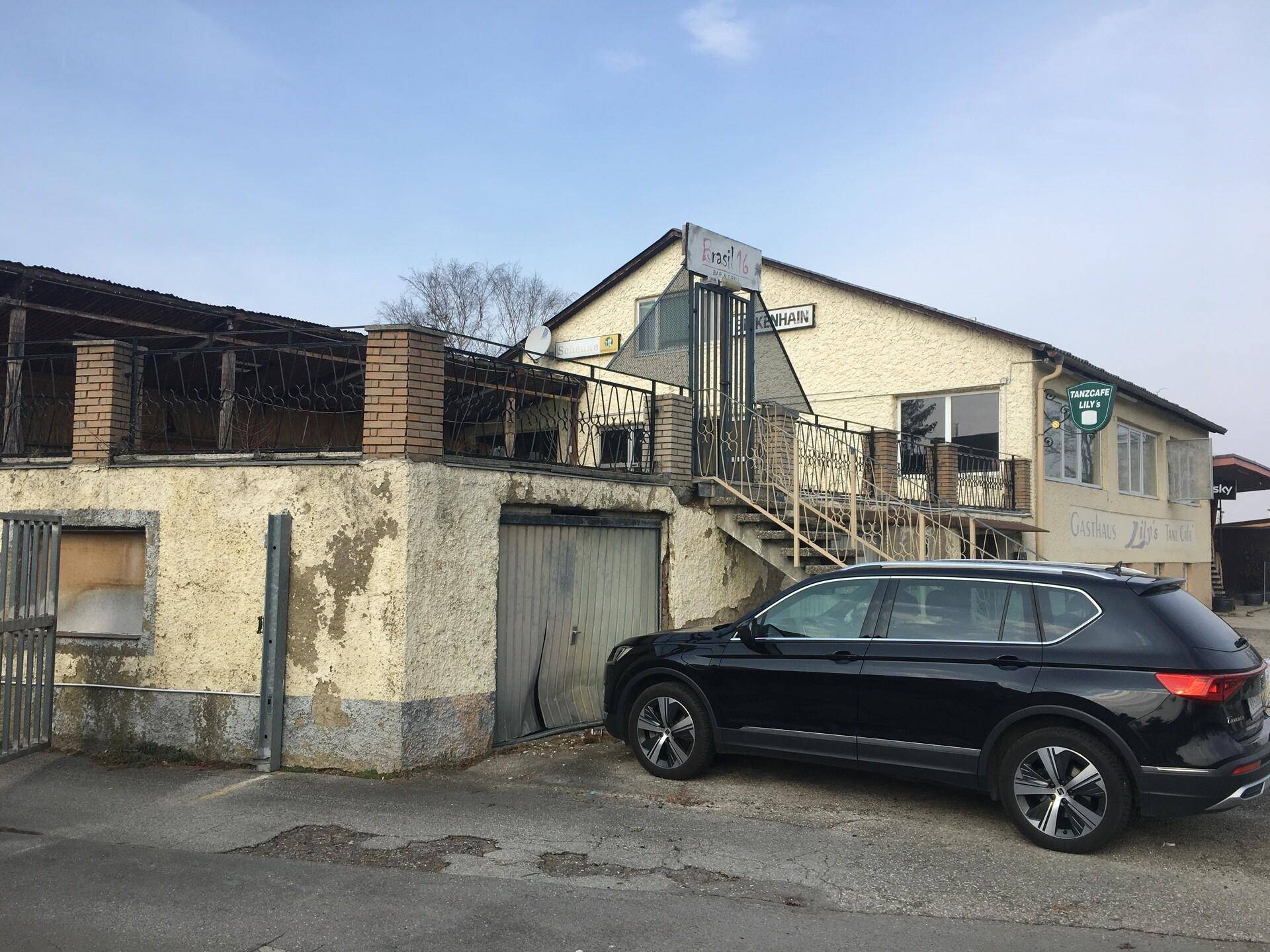 Gebäudeansicht eines Gasthauses mit einem schwarzen SUV davor, zeigt eine Putzfassade und einen oberen Balkonbereich.