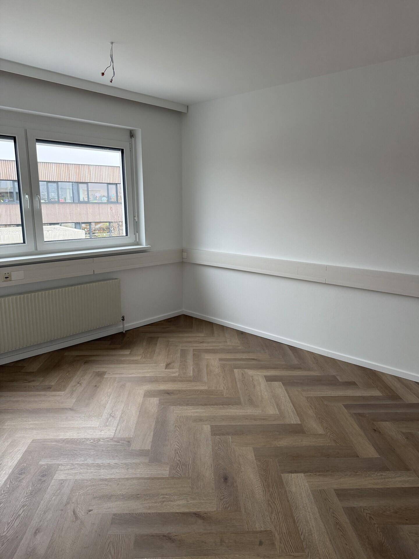 Heller, leerer Büroraum mit Fischgrätparkett, einem Fenster und einem Heizkörper an der Wand.