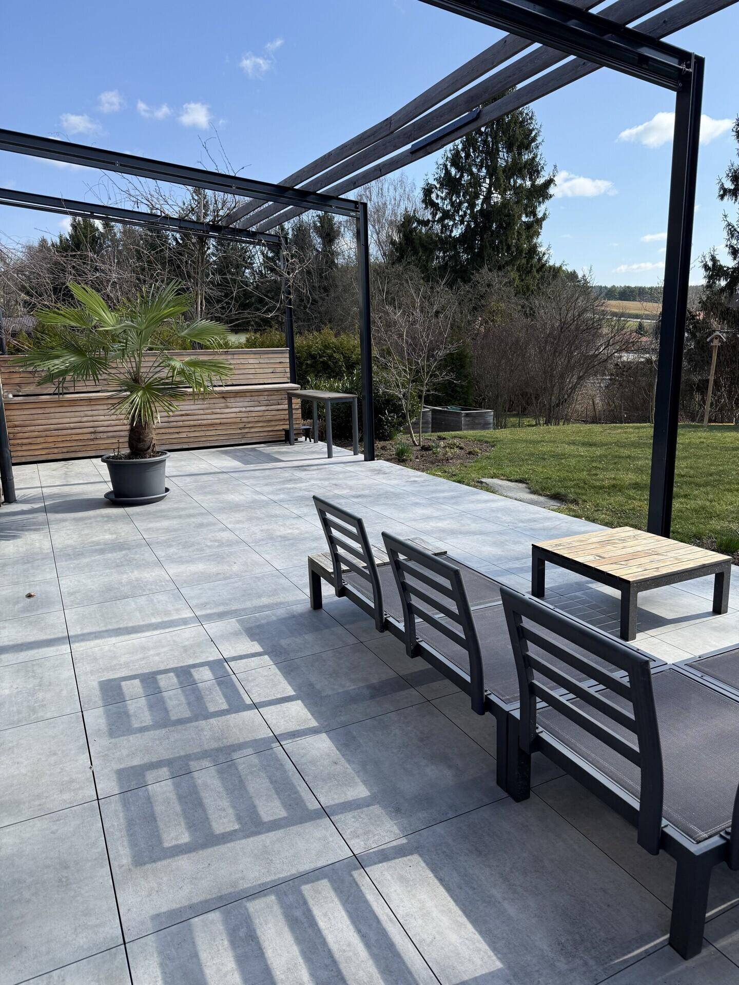 Moderne geflieste Terrasse mit Pergola, Outdoor-Möbeln und Blick ins Grüne unter blauem Himmel.