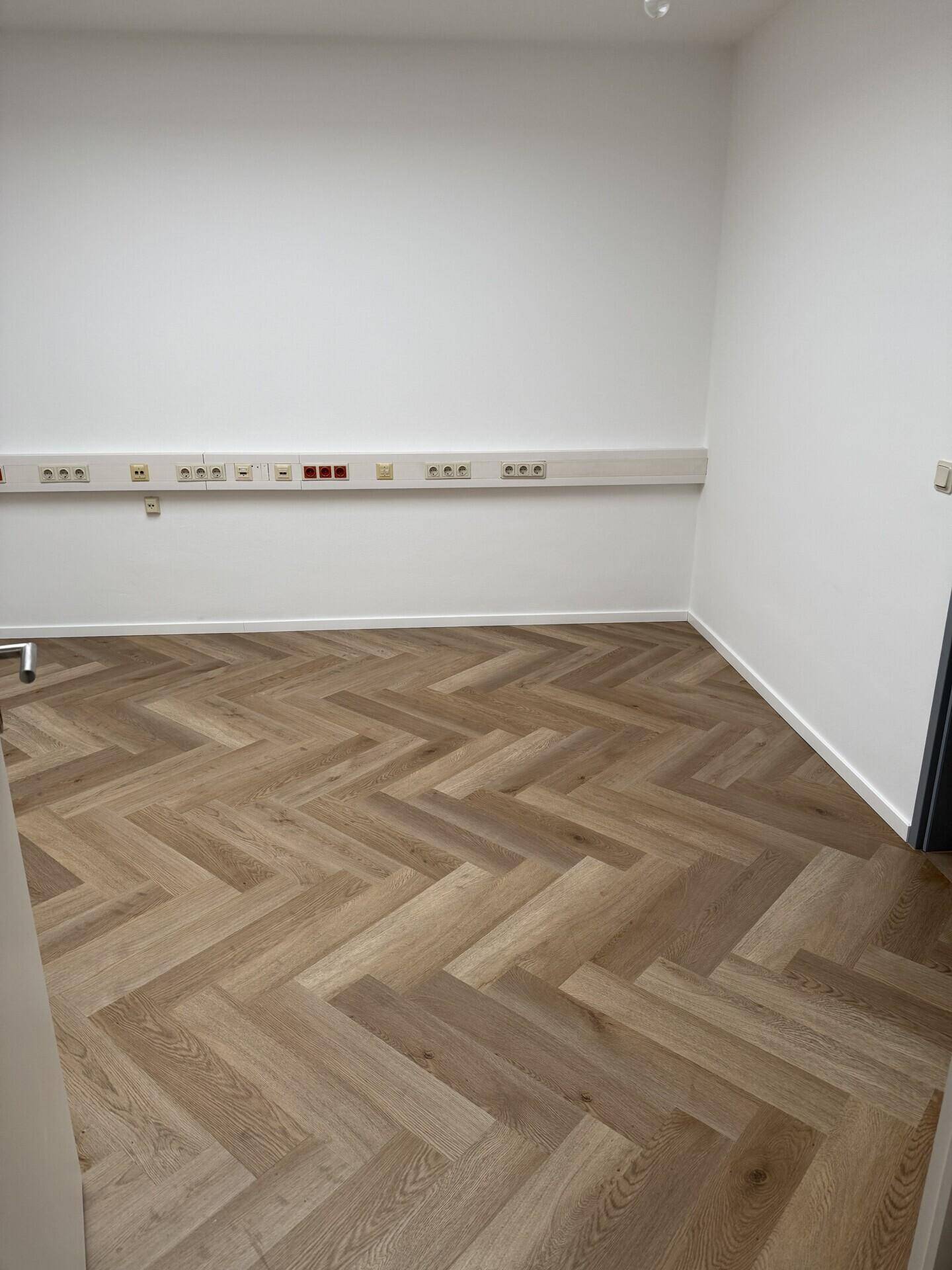 Leerer Büroraum mit modernem Fischgrätparkett und zahlreichen Steckdosen entlang der Wand.