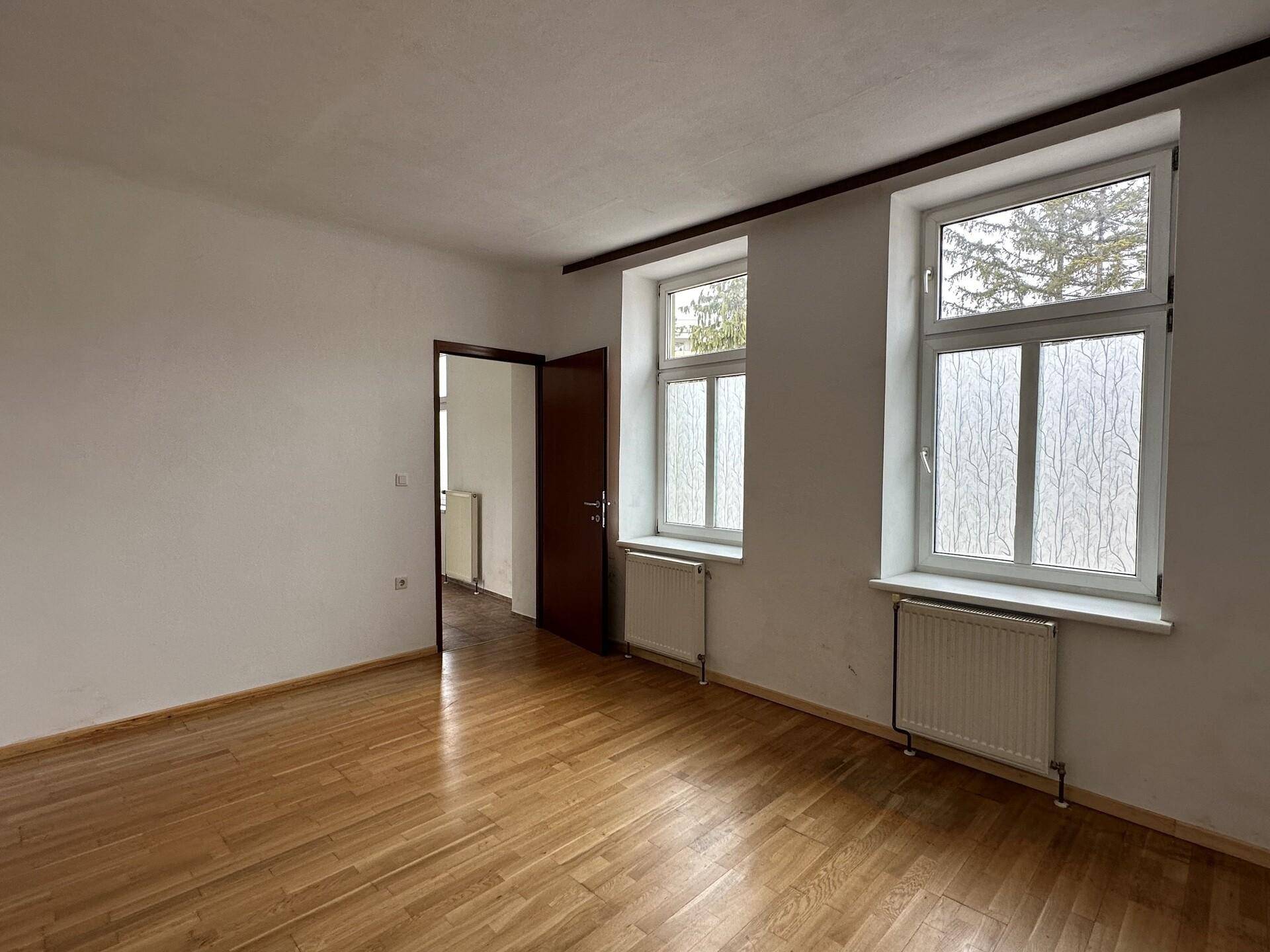 Geräumiges Wohnzimmer mit Holzparkettboden und zwei großen Fenstern, die viel Licht hereinlassen.