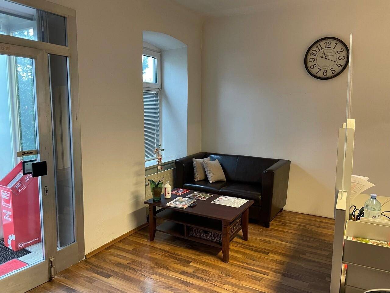 Gemütlicher Wartebereich mit dunklem Sofa, Couchtisch und Holzboden neben dem Eingangsbereich.