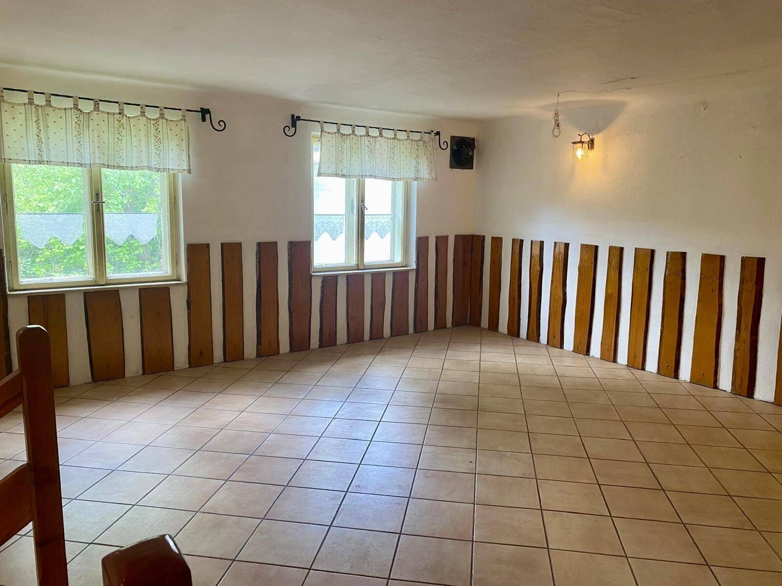 Einfacher Raum mit Fliesenboden, Holzvertäfelung und zwei Fenstern mit Blick ins Grüne.