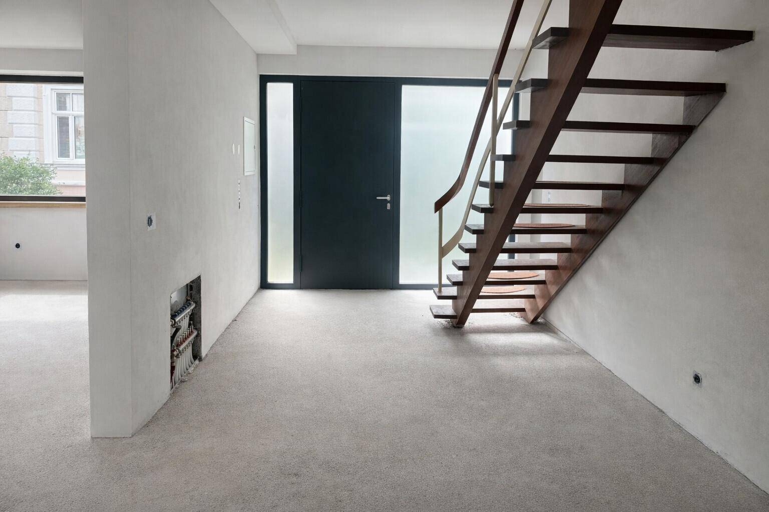 Heller Eingangsbereich im Rohbau mit moderner Holztreppe und großem Fenster.