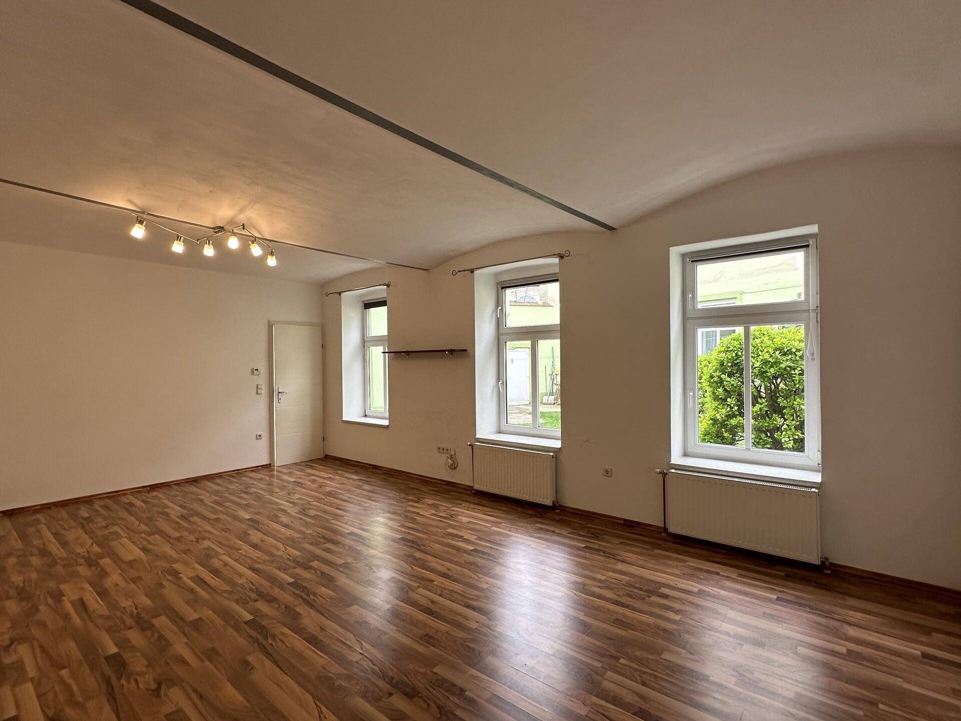 Leeres Wohnzimmer mit Holzboden und Blick auf den grünen Außenbereich.