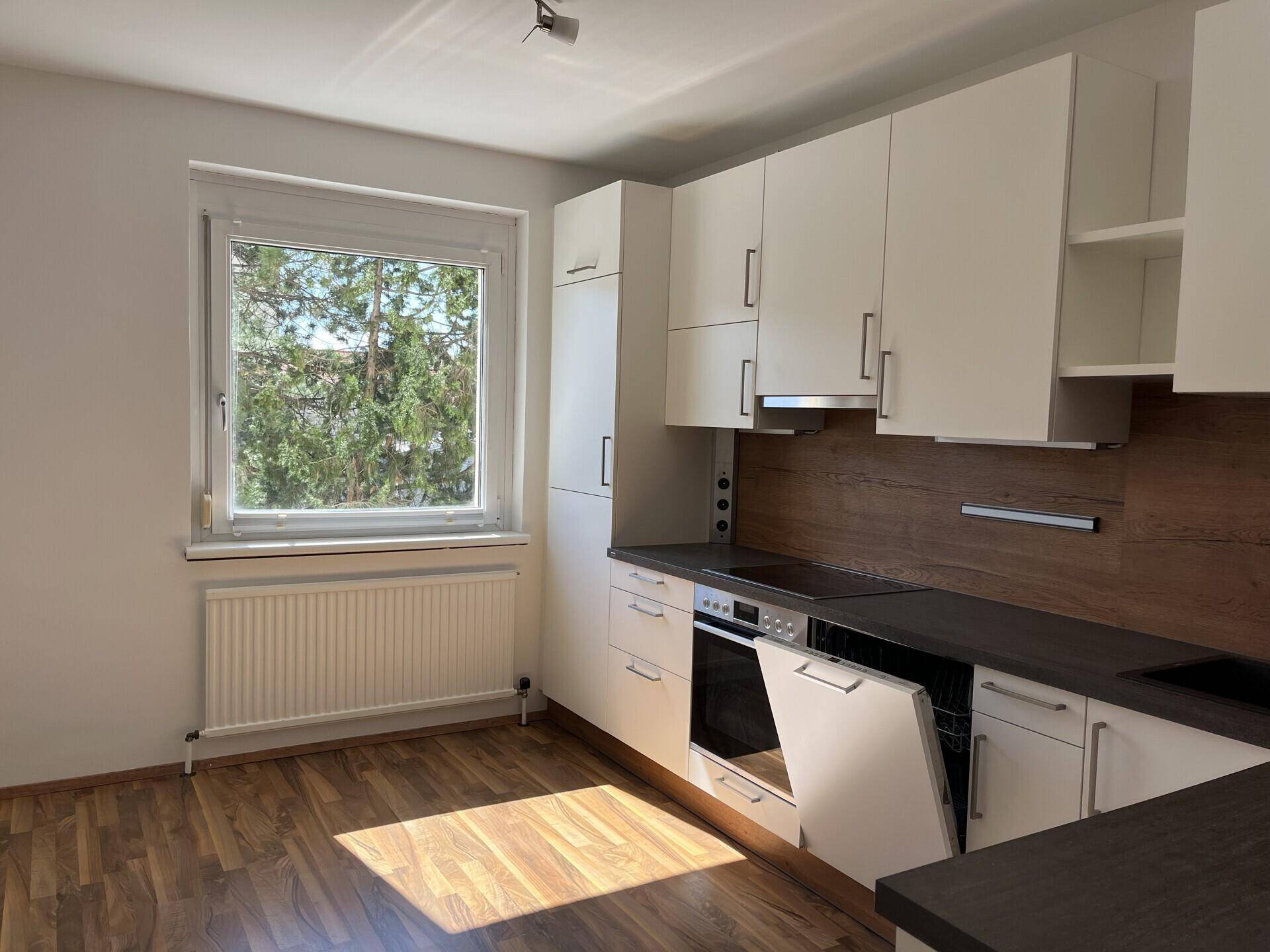 Moderne Einbauküche mit weißen Schränken, dunkler Arbeitsplatte und Fenster mit Grünblick.