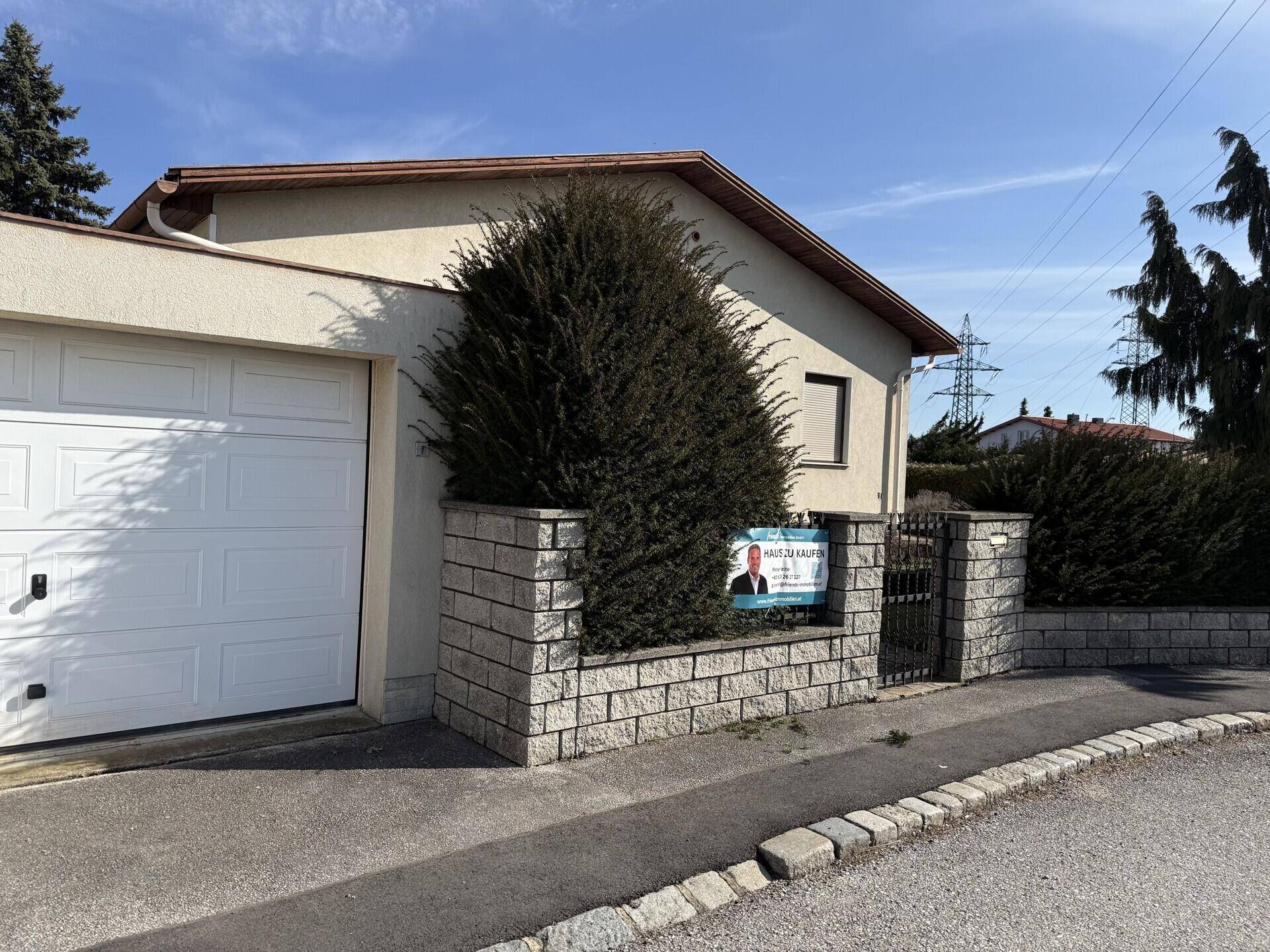 Hausfront mit Garage, Hecke und einem 'Haus zu Kaufen' Schild, einladend gestaltet.