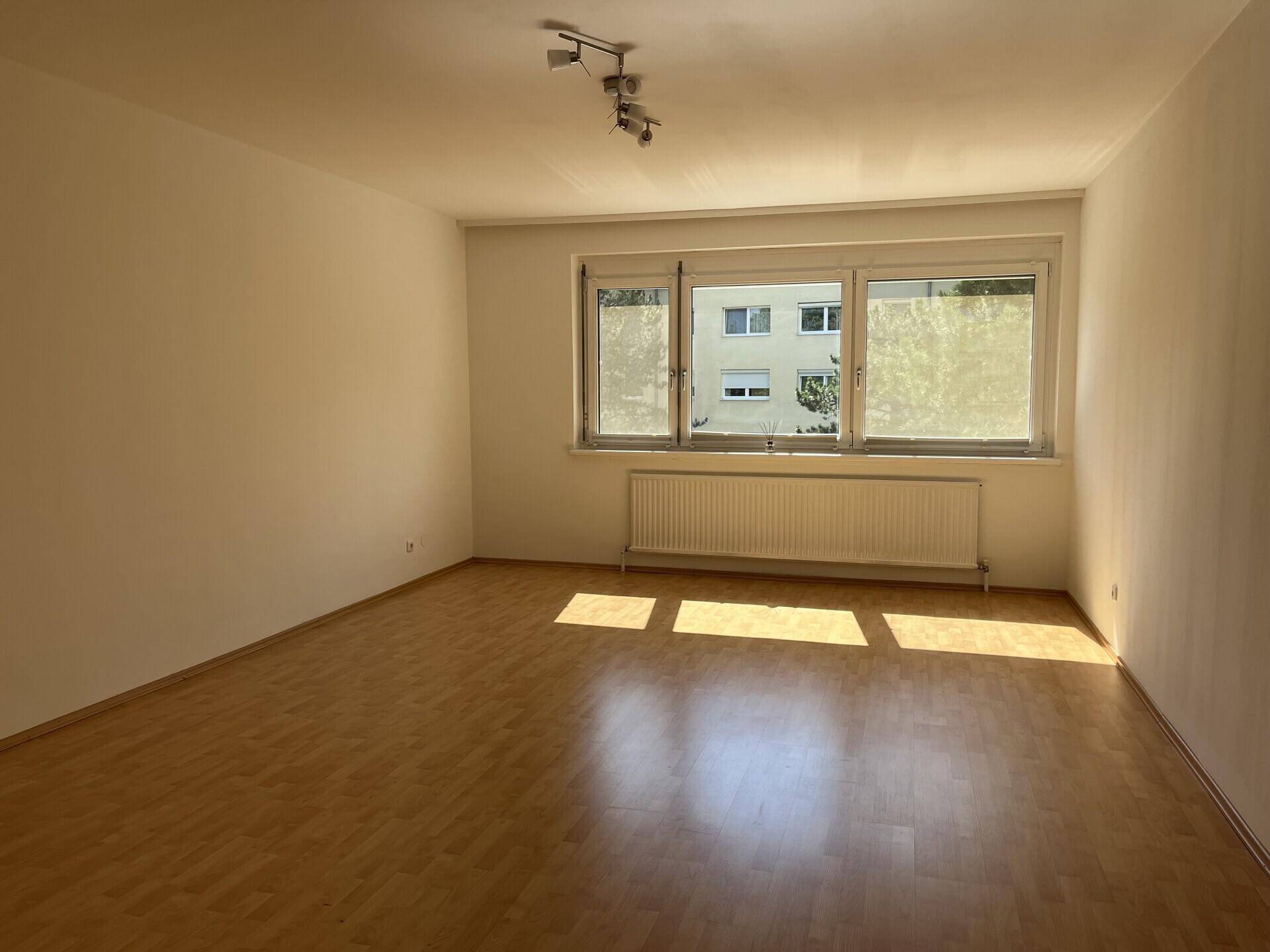 Helles Wohnzimmer mit Laminatboden und großem Fenster, das viel Tageslicht hereinlässt.