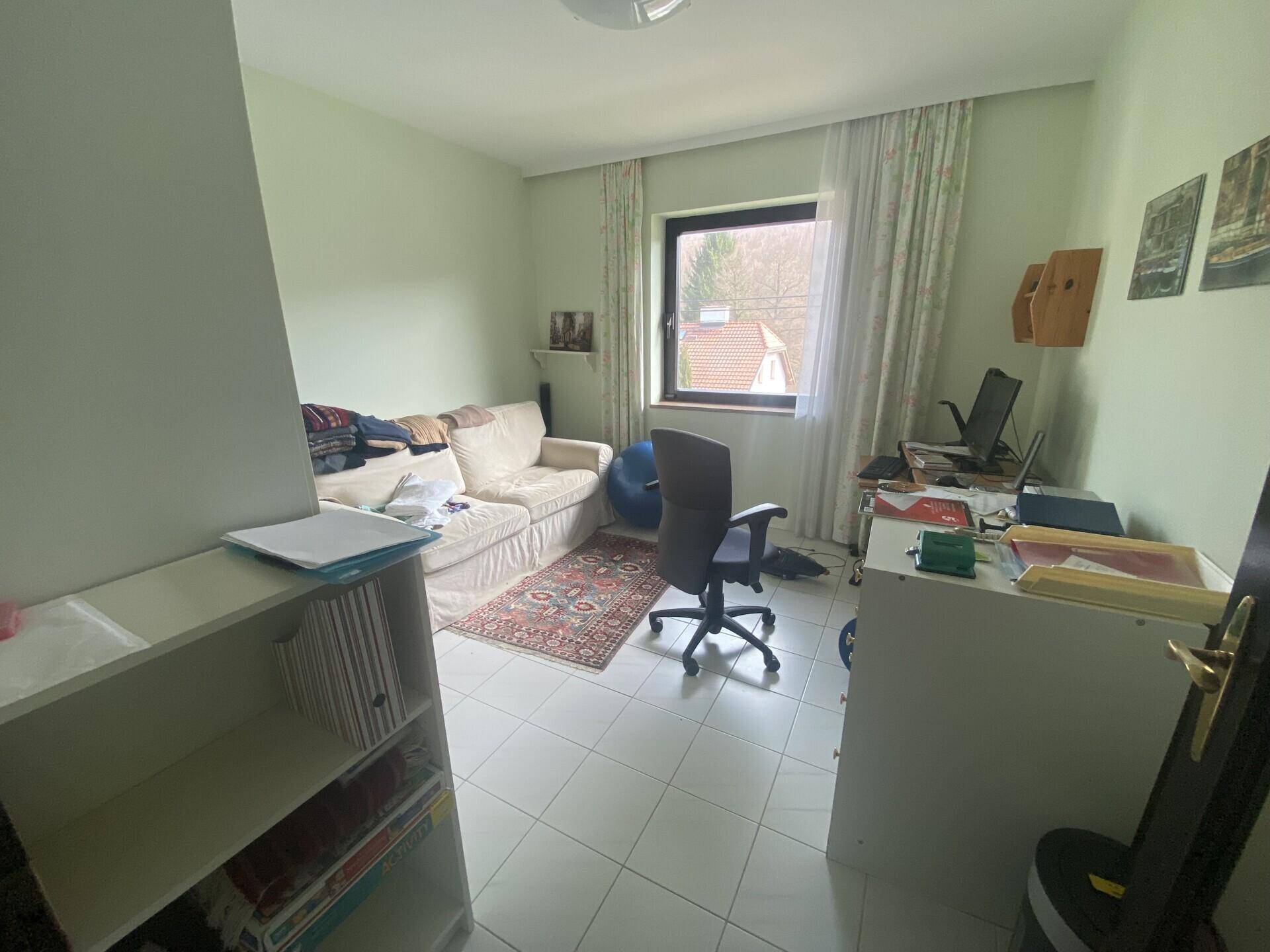 Helles Zimmer mit Fliesenboden, Sofa und Schreibtisch, nutzbar als Schlaf- oder Arbeitszimmer.
