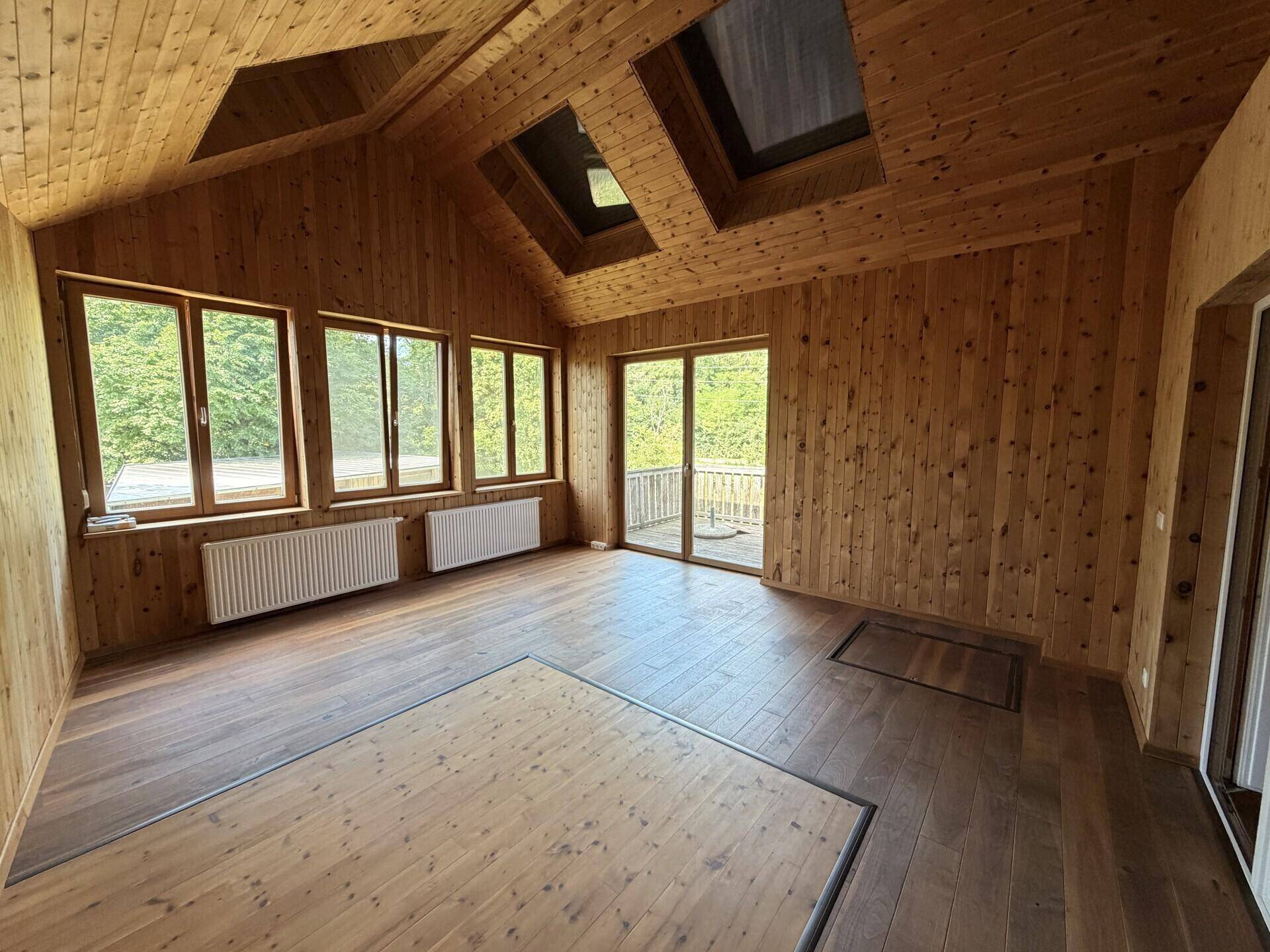 Geräumiger Raum mit Holzverkleidung an Wänden und Decke, großen Fenstern und Zugang zum Balkon.