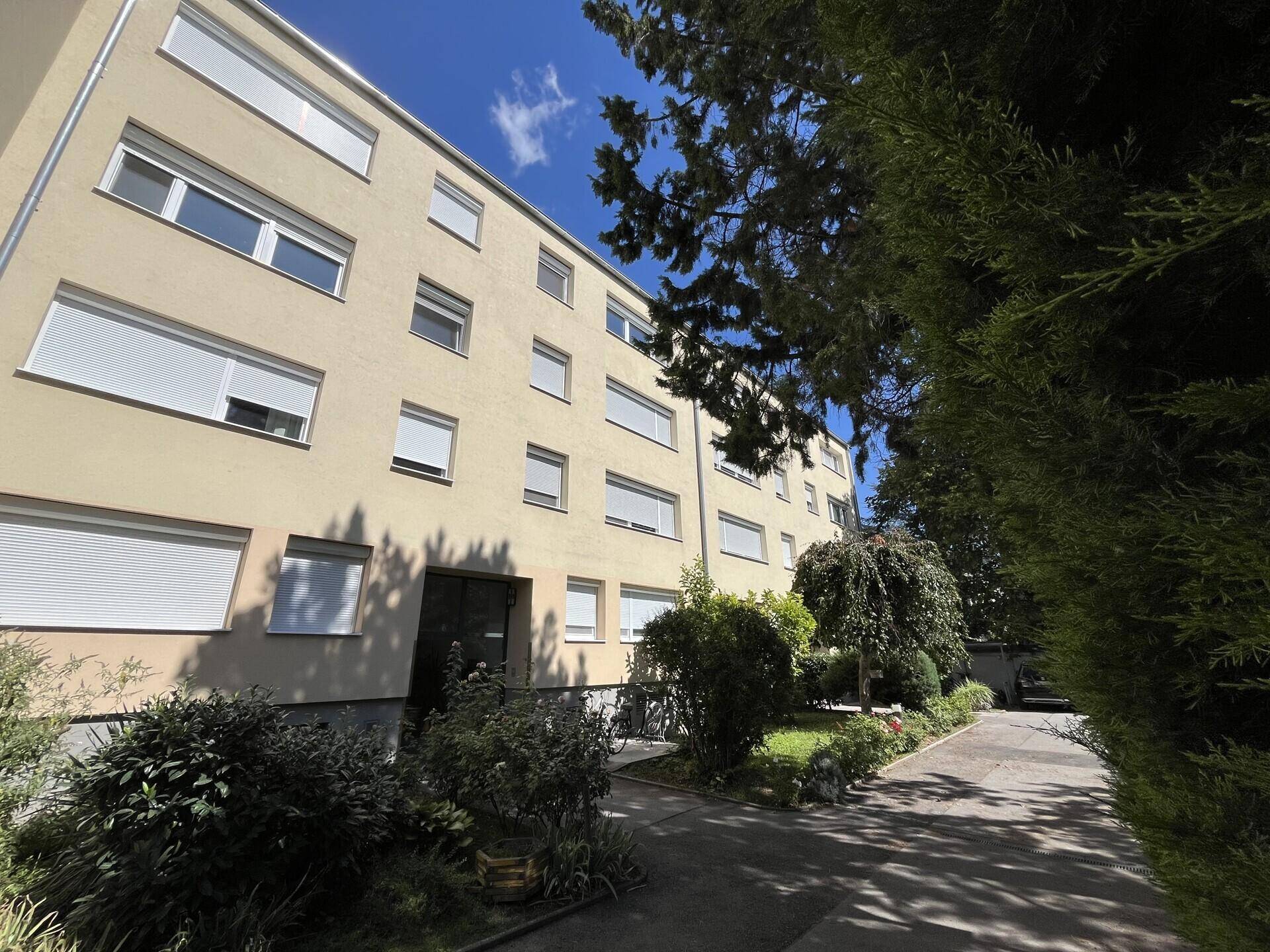 Mehrfamilienhaus mit heller Fassade, umgeben von Bäumen und einem gepflegten Vorgarten.