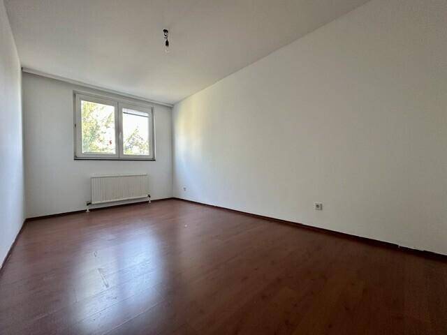 Leeres Schlafzimmer mit Laminatboden, weißen Wänden und einem Fenster, das viel Tageslicht hereinlässt.