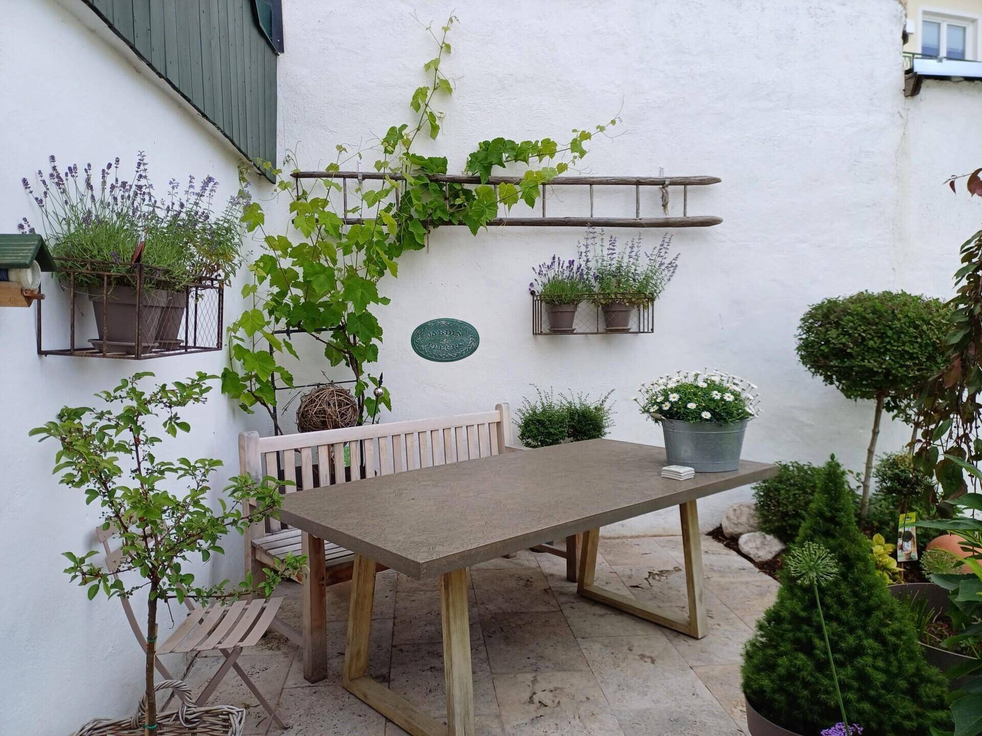 Gemütlicher Innenhof mit Steinfliesen, Holztisch und Bank, umgeben von Pflanzen.