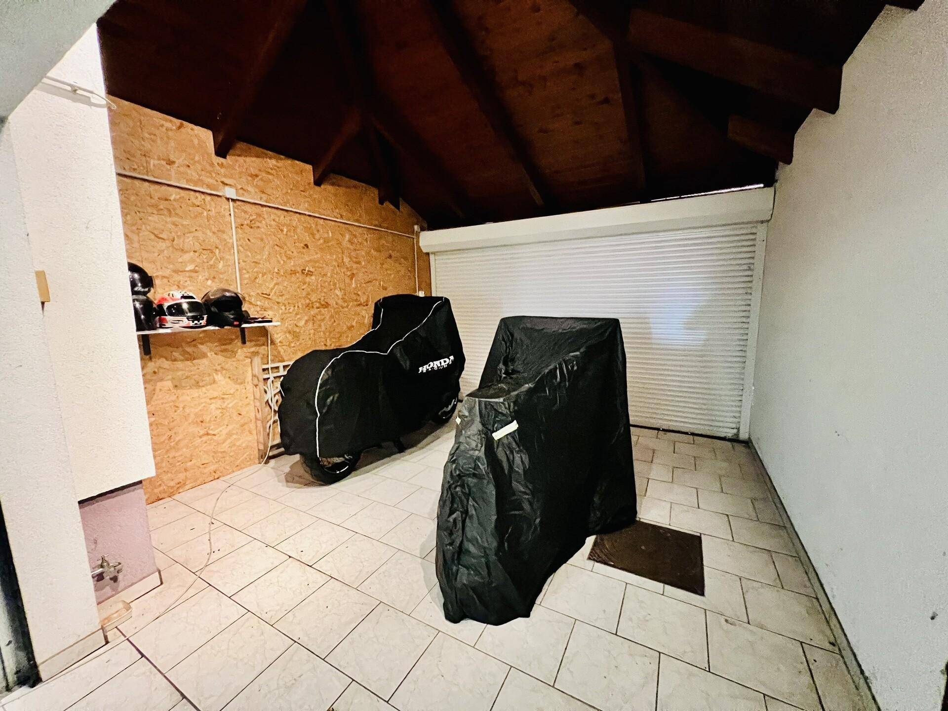 Abstellbereich in der Garage mit Fliesenboden, Holzdecke und Platz für Motorräder hinter einem Rolltor.