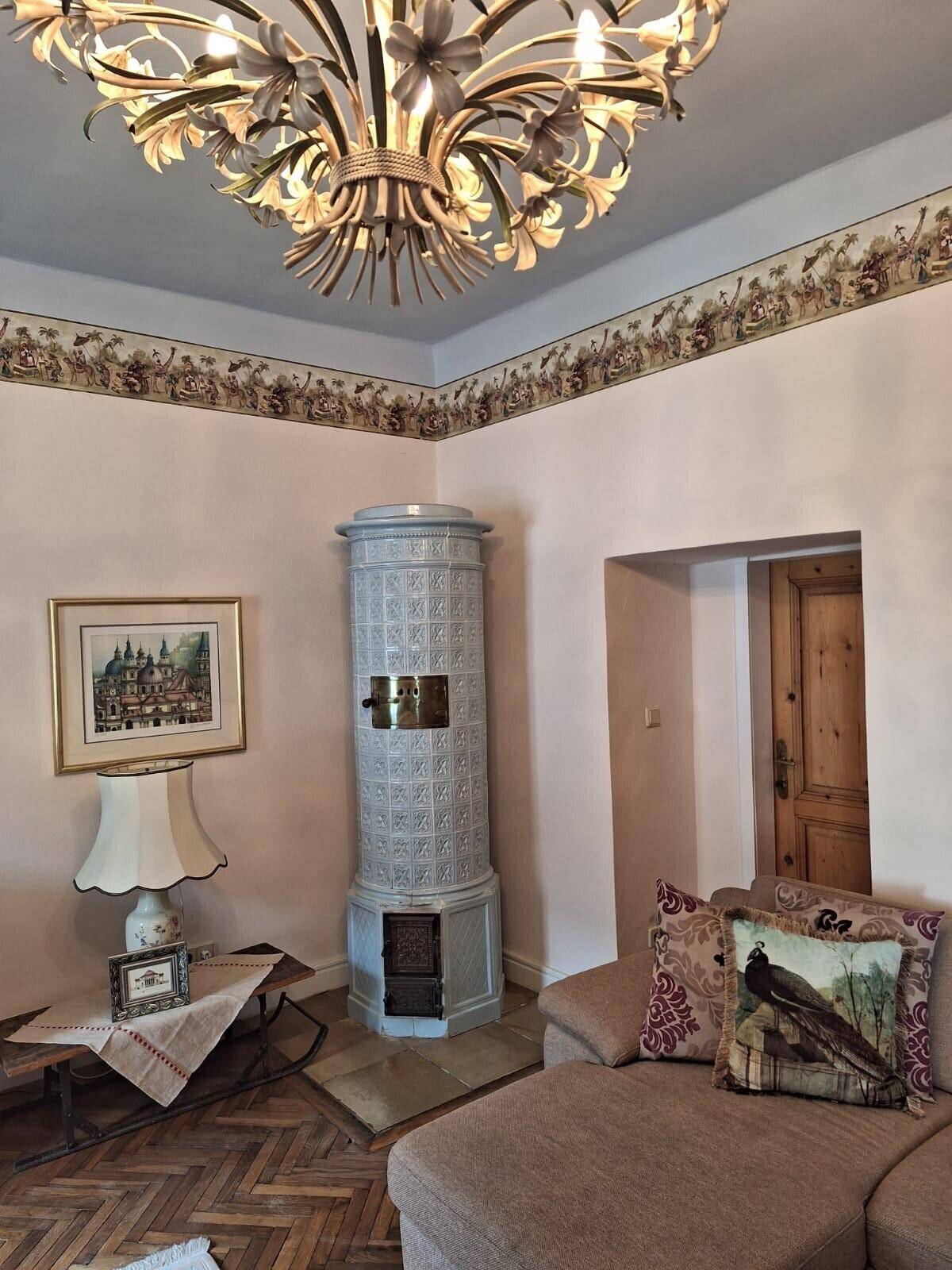 Gemütliches Wohnzimmer mit Kachelofen, Sofa und floraler Bordüre an der Wand.