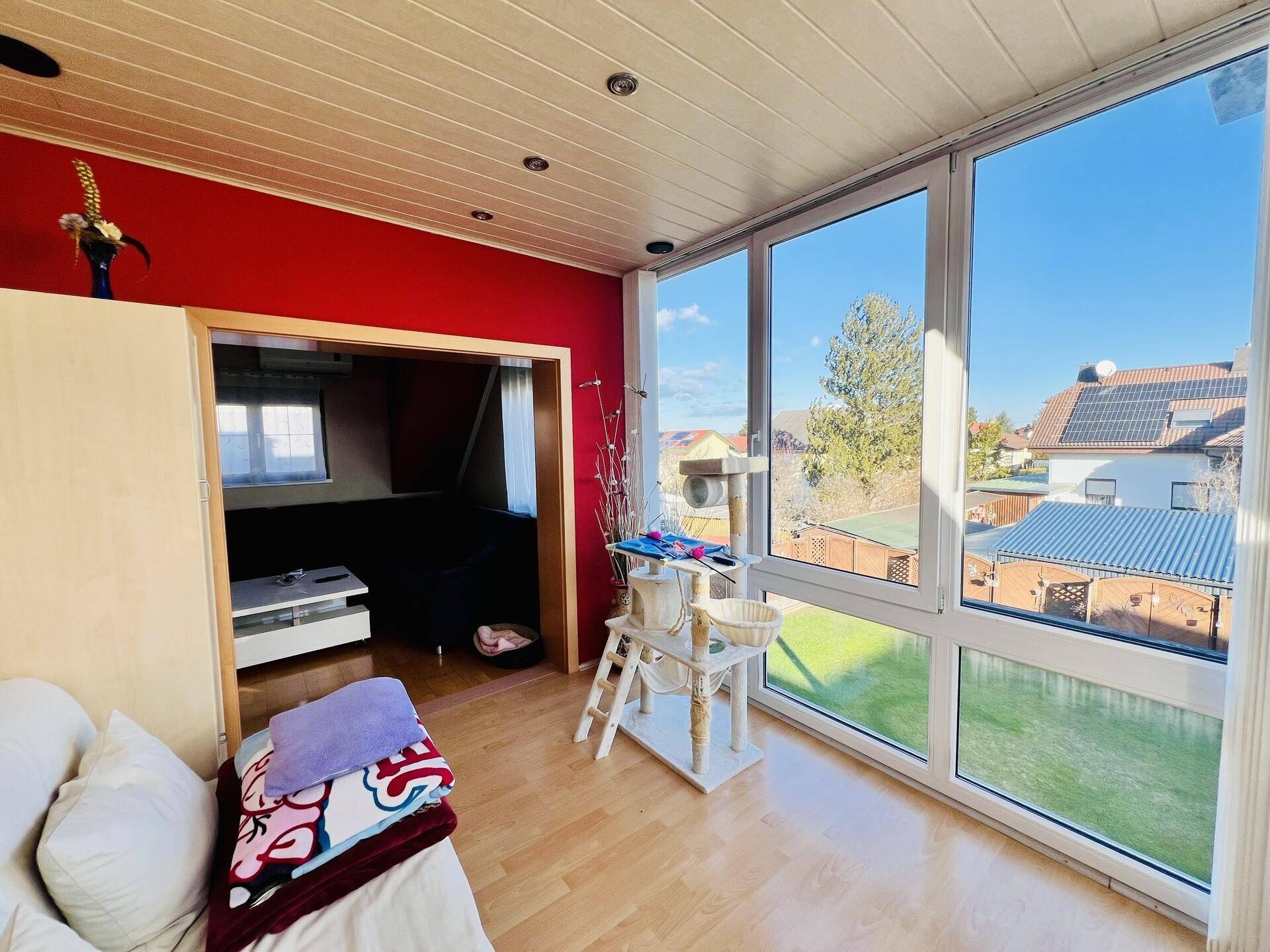 Lichtdurchfluteter Raum mit großen Fenstern, Holzboden und Zugang zu einem weiteren Zimmer.