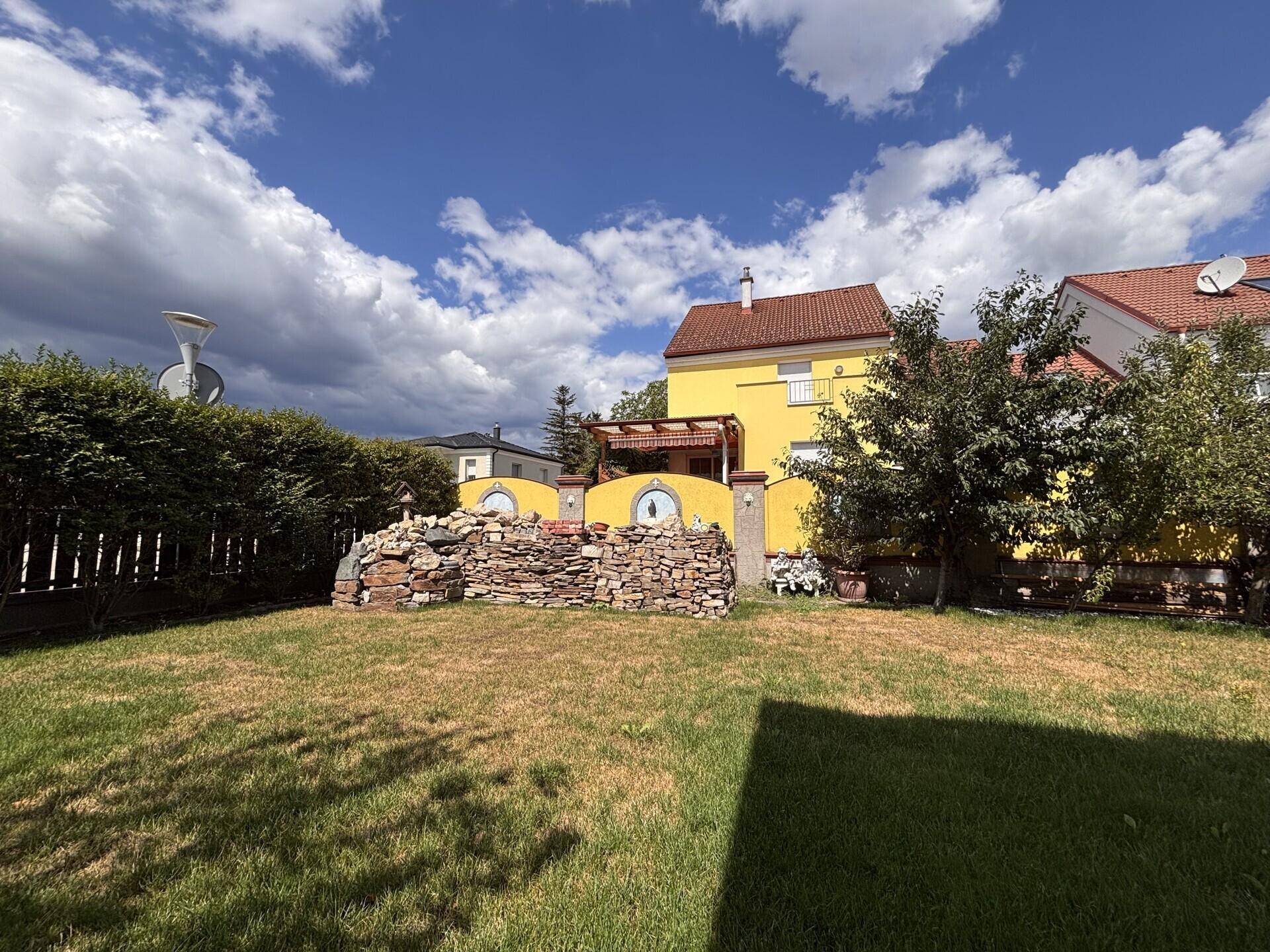 Gepflegter Garten mit Rasenfläche, Hecke und einem gelben Haus im Hintergrund.