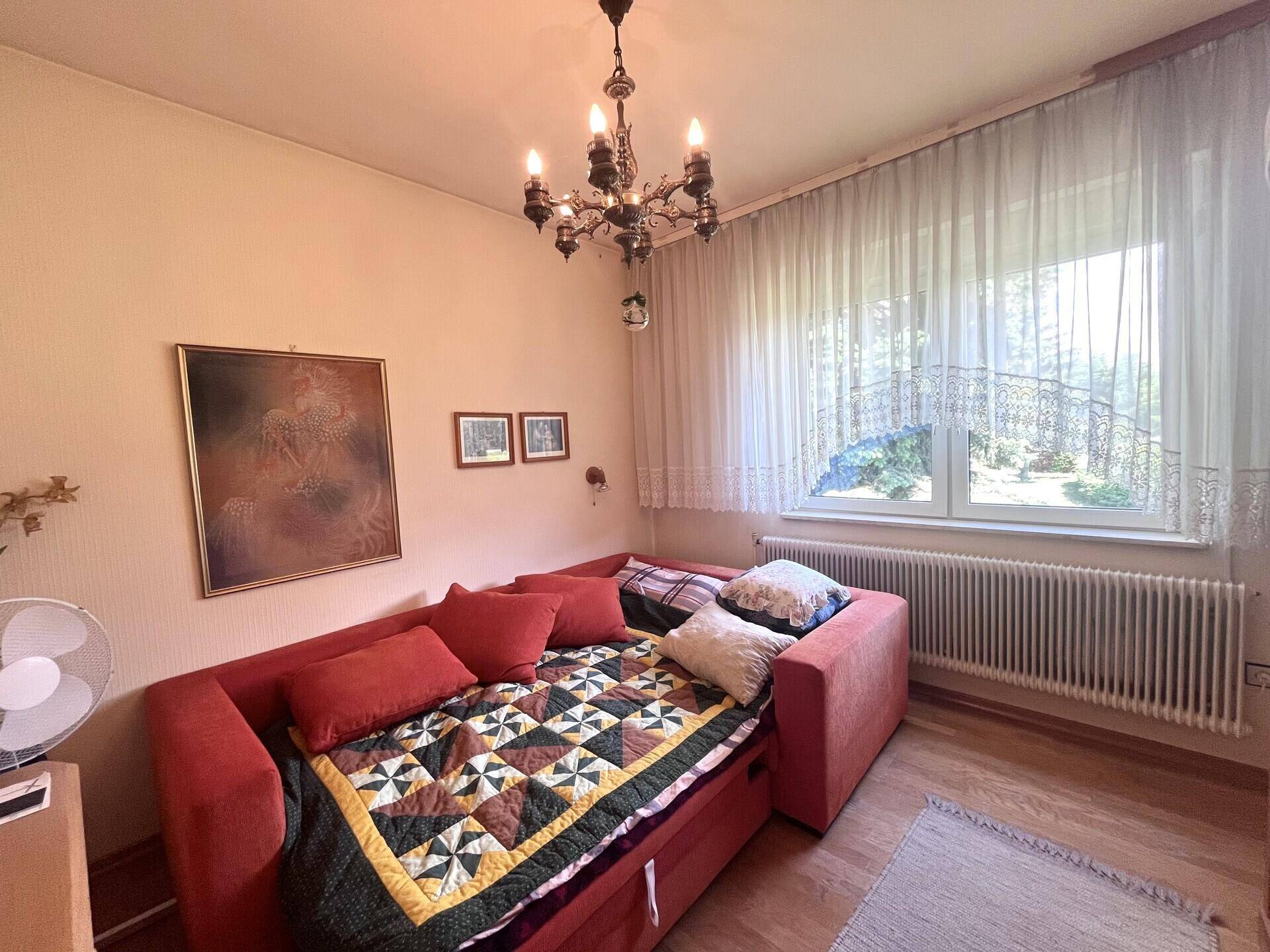 Helles Schlafzimmer mit Tagesbett, großem Fenster und klassischem Kronleuchter, ideal als Gästezimmer.