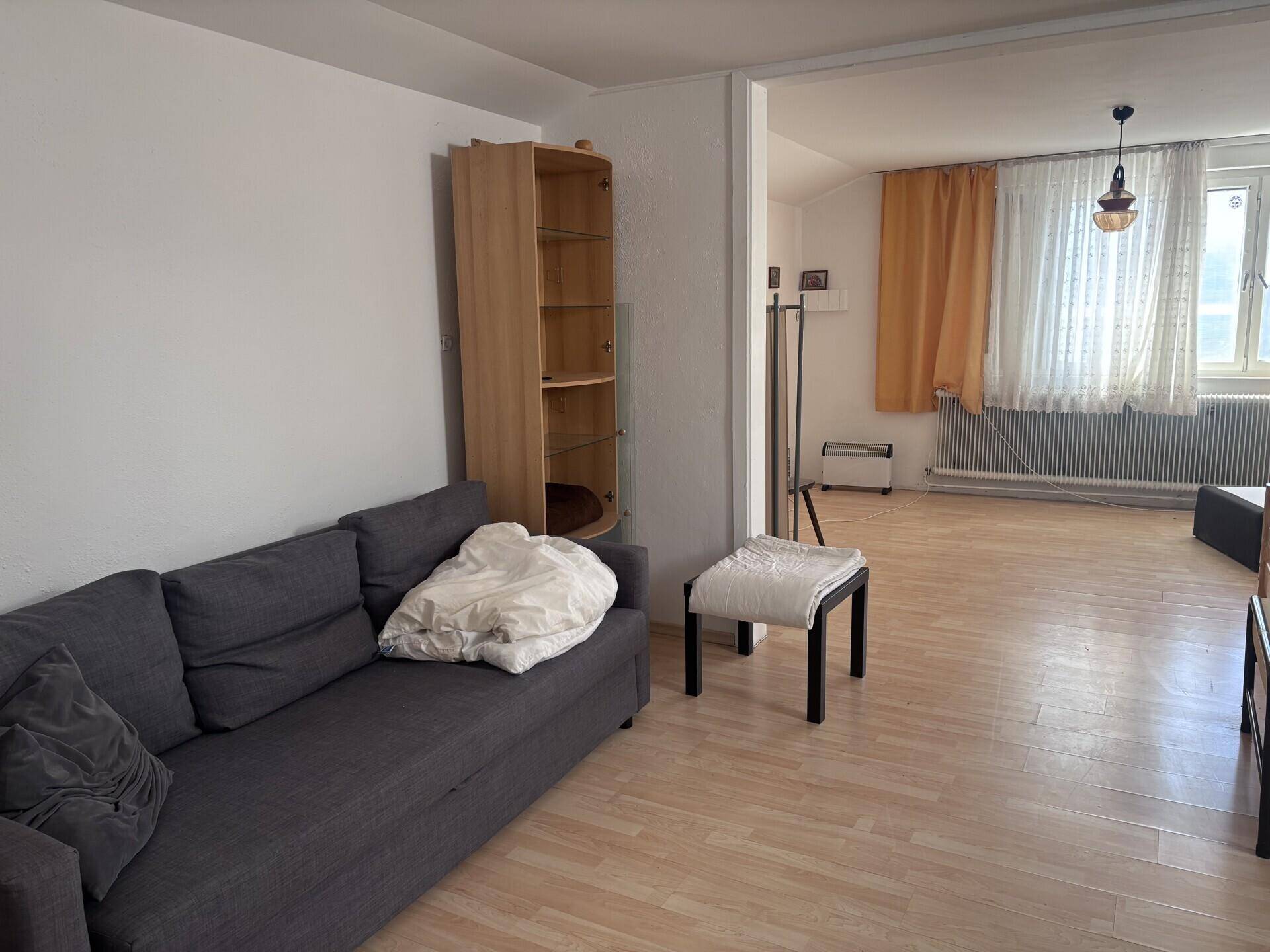 Heller Wohnbereich mit einem grauen Sofa, einem Holzregal und hellem Laminatboden, gut beleuchtet durch Fenster.