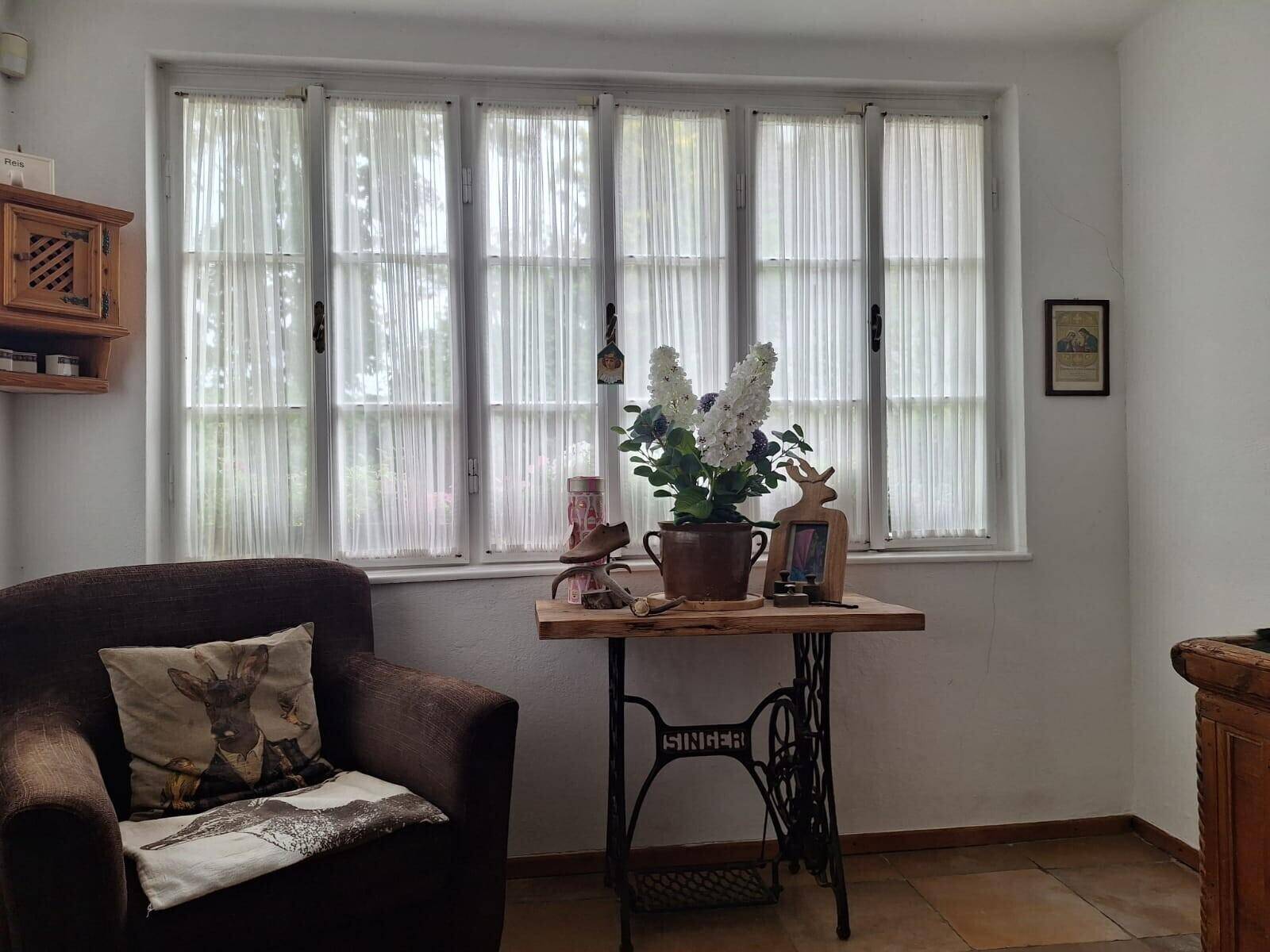 Helles Zimmer mit großen Fenstern, Vorhängen und einem bequemen Sessel.