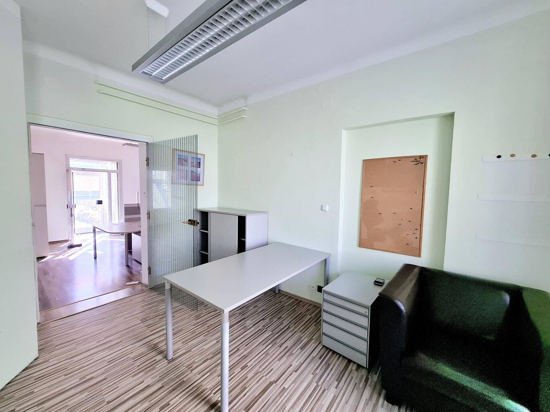 Helles Büro mit hellgrünen Wänden, Holzboden und einem Schreibtisch, der zu einem weiteren Raum führt.