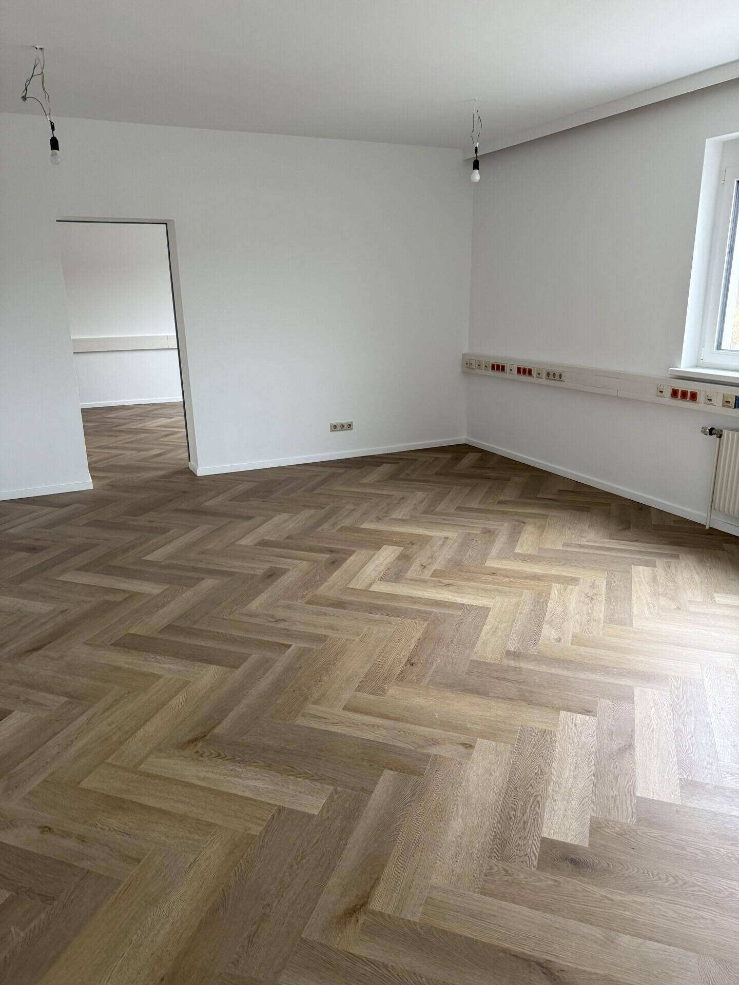 Leerer Büroraum mit modernem Fischgrätparkett und zahlreichen Steckdosen entlang der Wand.