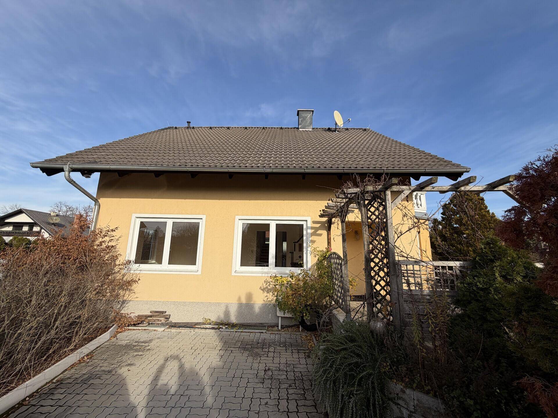 Vorderansicht des Hauses mit gepflasterter Einfahrt, Pergola und gepflegter Bepflanzung.