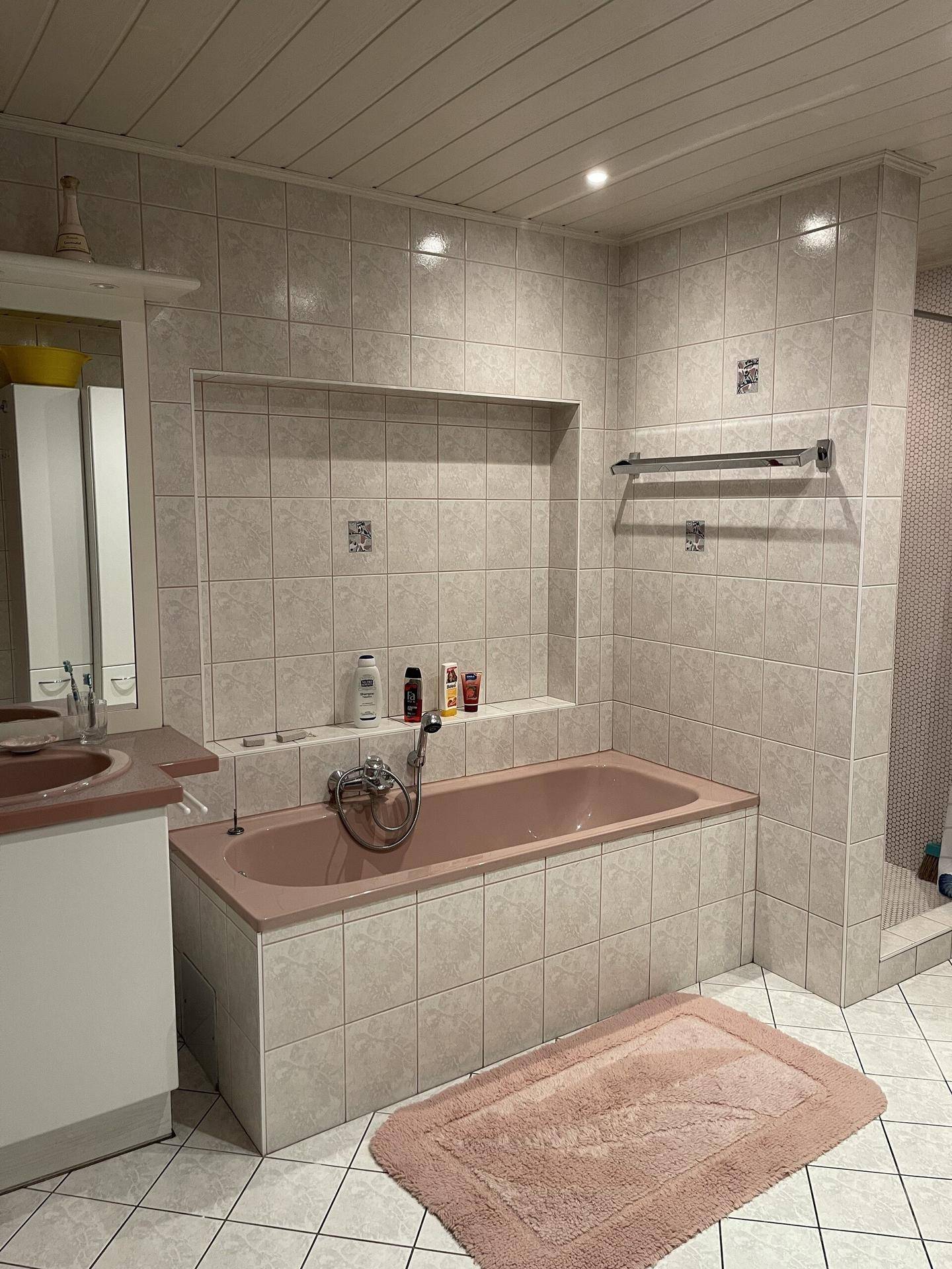 Badezimmer mit rosa Badewanne, hellen Fliesen und integrierter Ablagefläche.