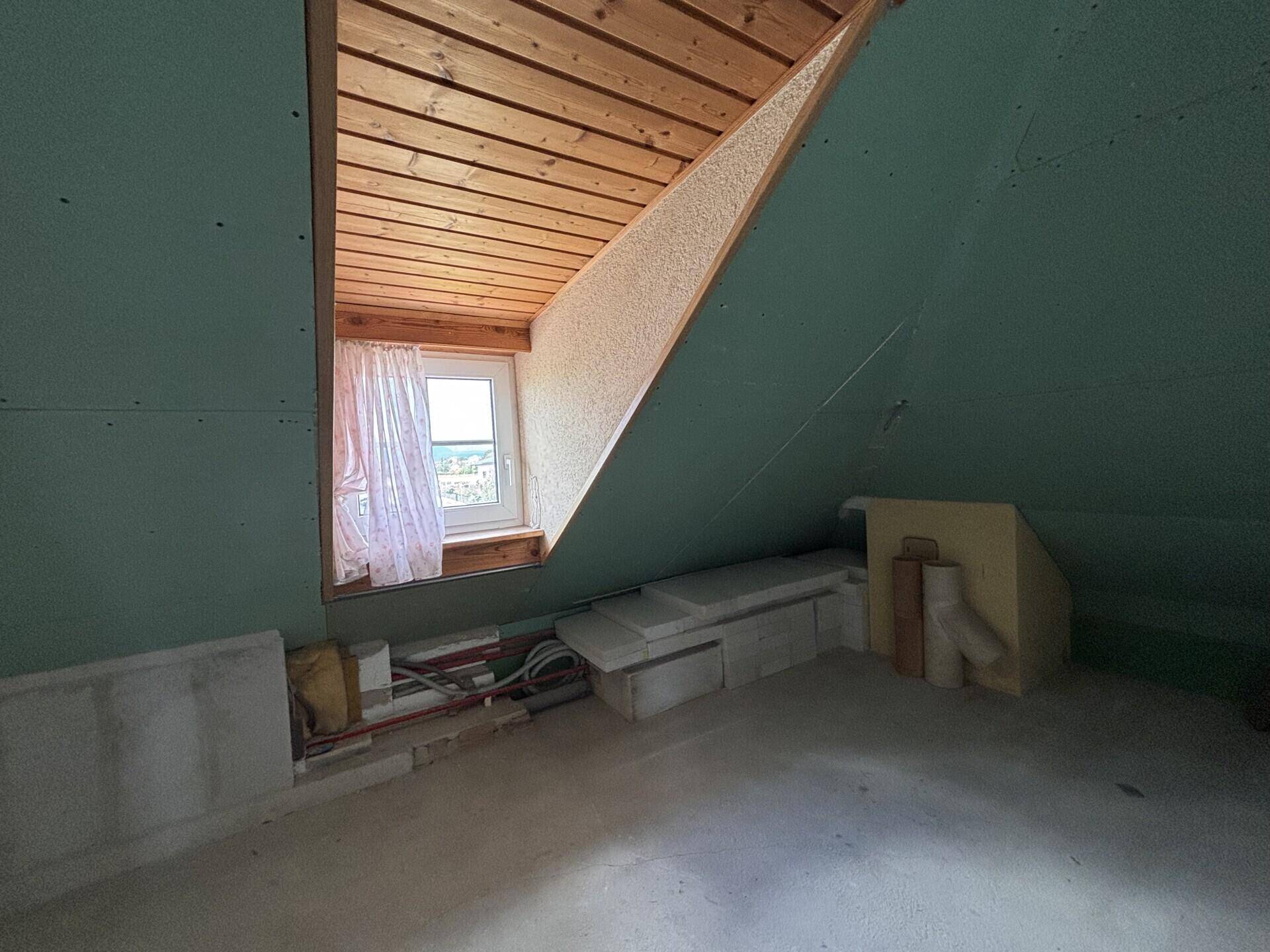 Dachgeschossraum im Rohbau mit schrägen Wänden und einem Dachfenster.