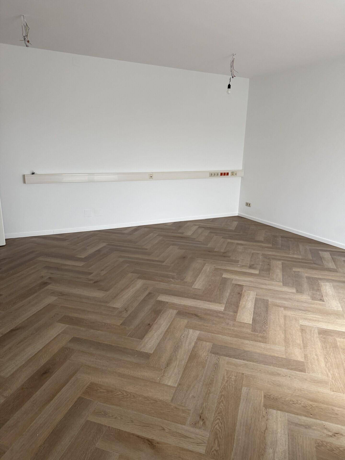Leerer Büroraum mit modernem Fischgrätparkett und zahlreichen Steckdosen entlang der Wand.