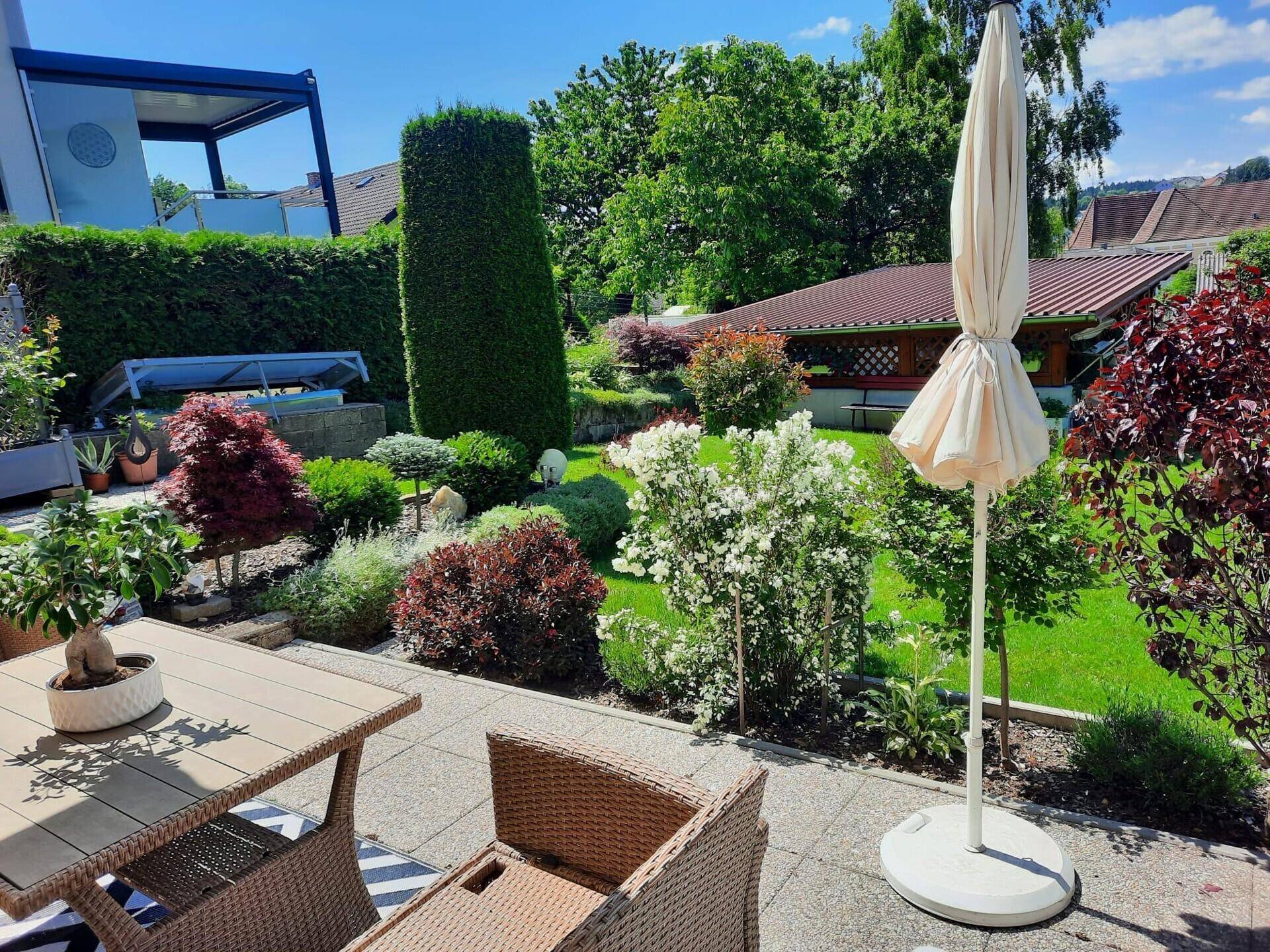 Terrasse mit Gartenmöbeln und Sonnenschirm, umgeben von üppiger Bepflanzung.