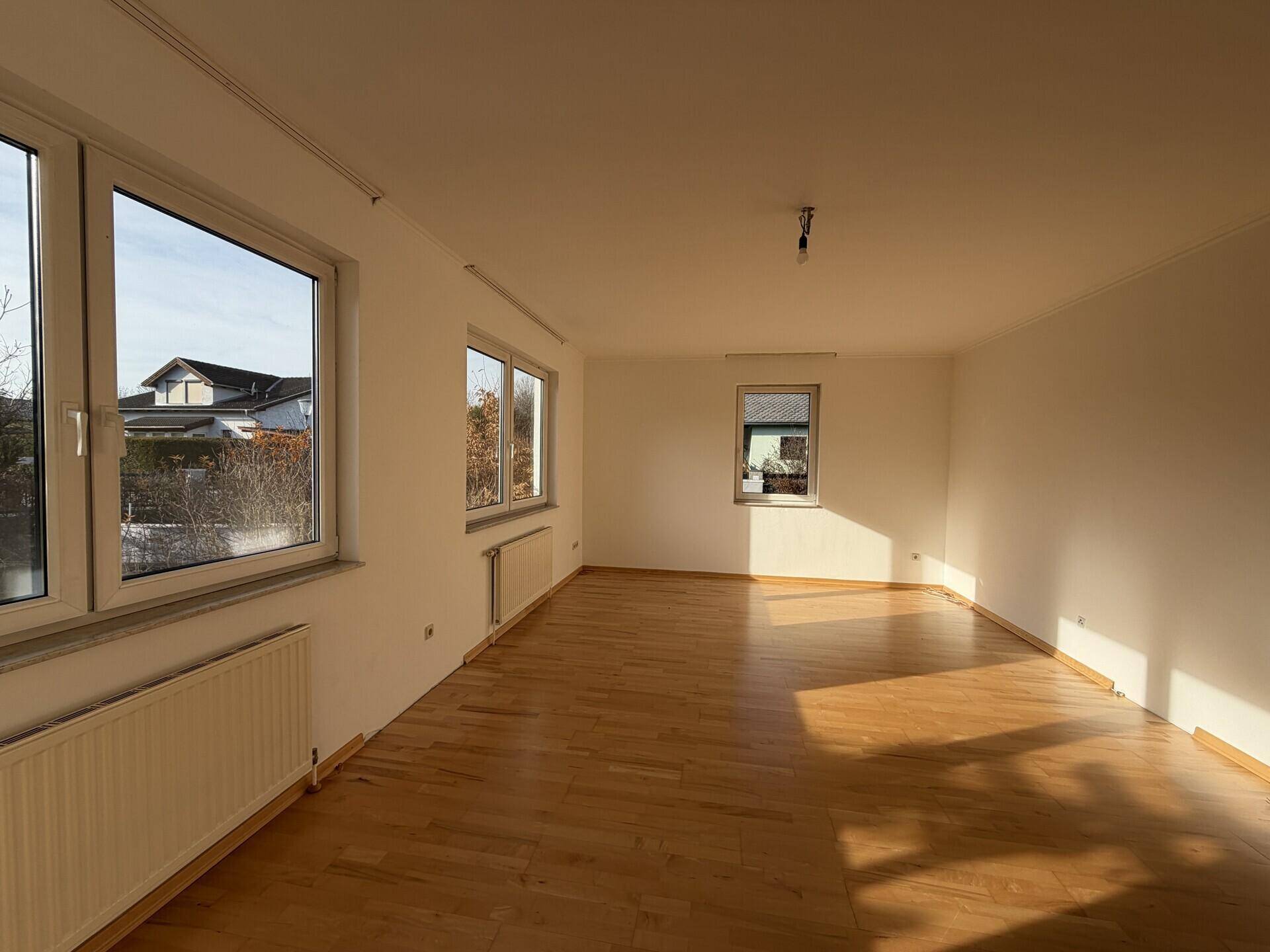 Geräumiger, heller Raum mit Parkettboden und mehreren Fenstern, die den Blick nach draußen freigeben.