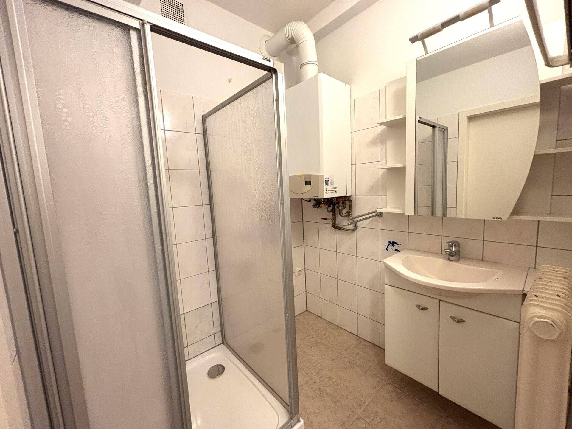 Badezimmer mit Duschkabine, Waschbecken und Boiler, hell gefliest und praktisch eingerichtet.