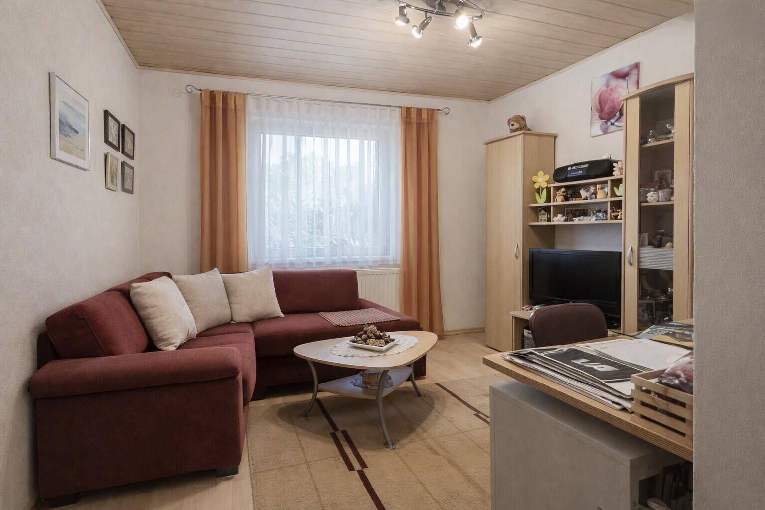 Helles Wohnzimmer mit Ecksofa, Teppichboden und einem Fenster mit Blick ins Grüne.