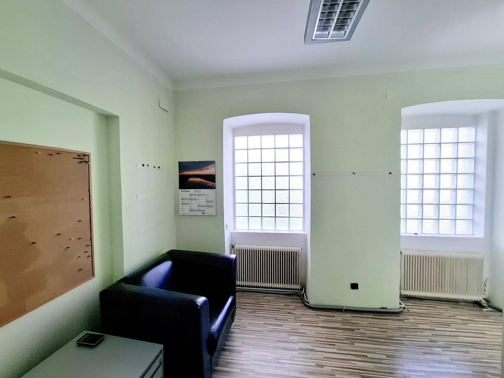 Büro mit hellgrünen Wänden, Holzboden und zwei Fenstern mit Glasbausteinen, ausgestattet mit einem Sessel.