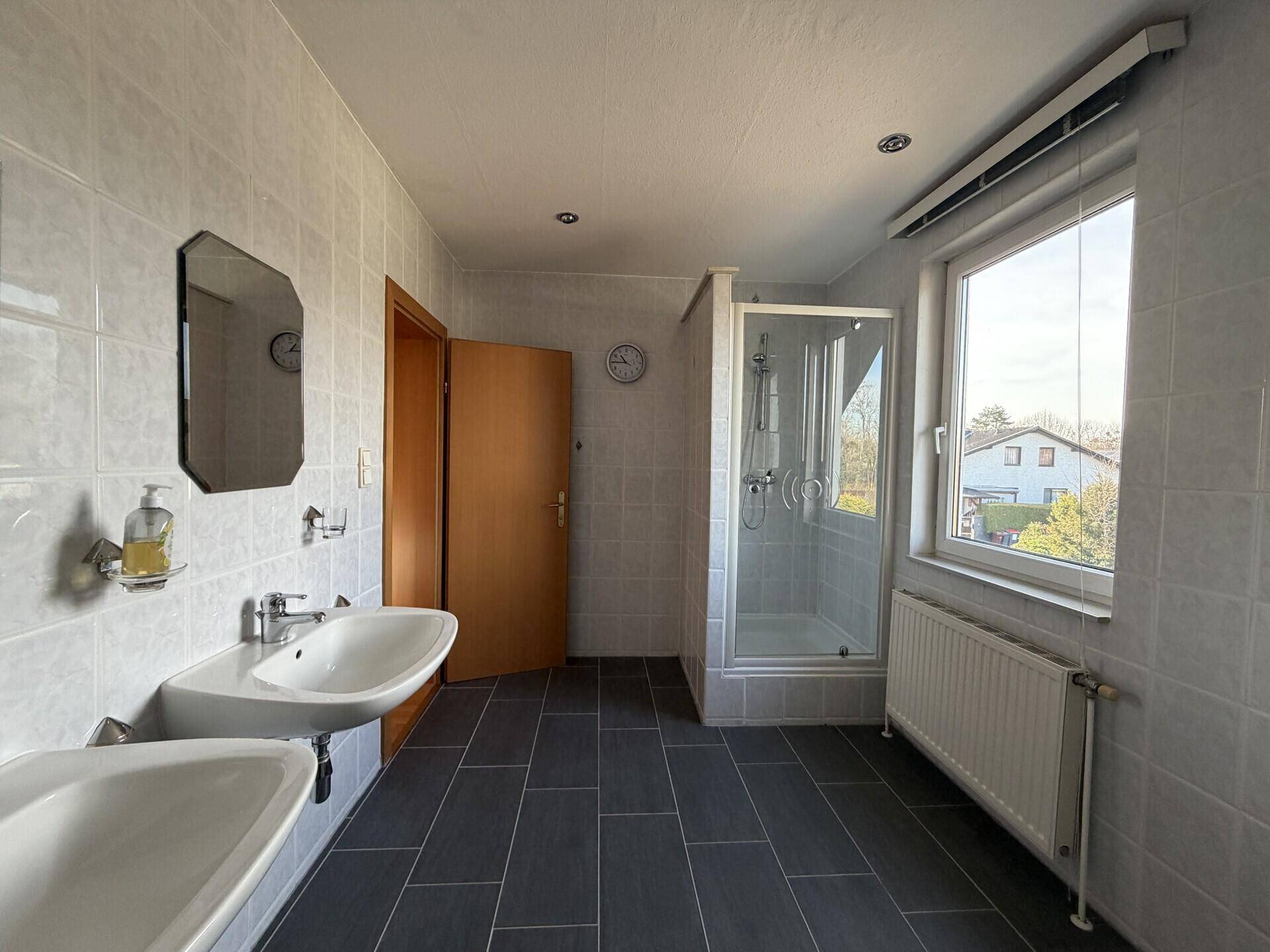 Geräumiges Badezimmer mit Doppelwaschbecken, Duschkabine und Fenster, hell gefliest.