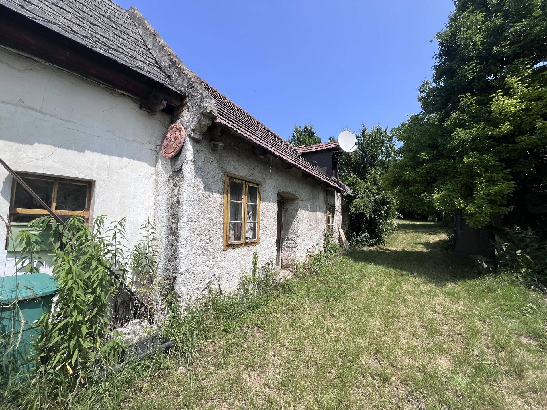 Altes Bauernhaus mit verwitterter Fassade und einem sonnigen, verwilderten Gartenbereich.