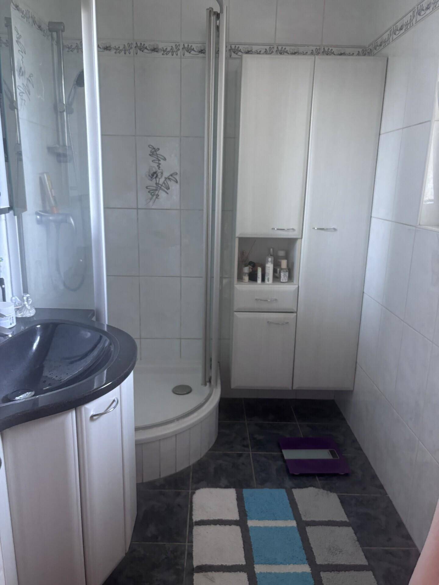 Badezimmer mit Duschkabine, dunklem Waschbecken und weißen Schränken, ausgestattet mit Fliesenboden.
