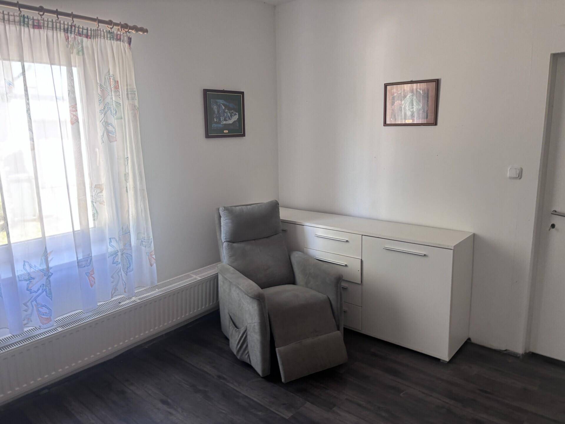 Wohnraum mit einem bequemen Sessel, einem weißen Sideboard und einem Fenster mit Vorhängen.