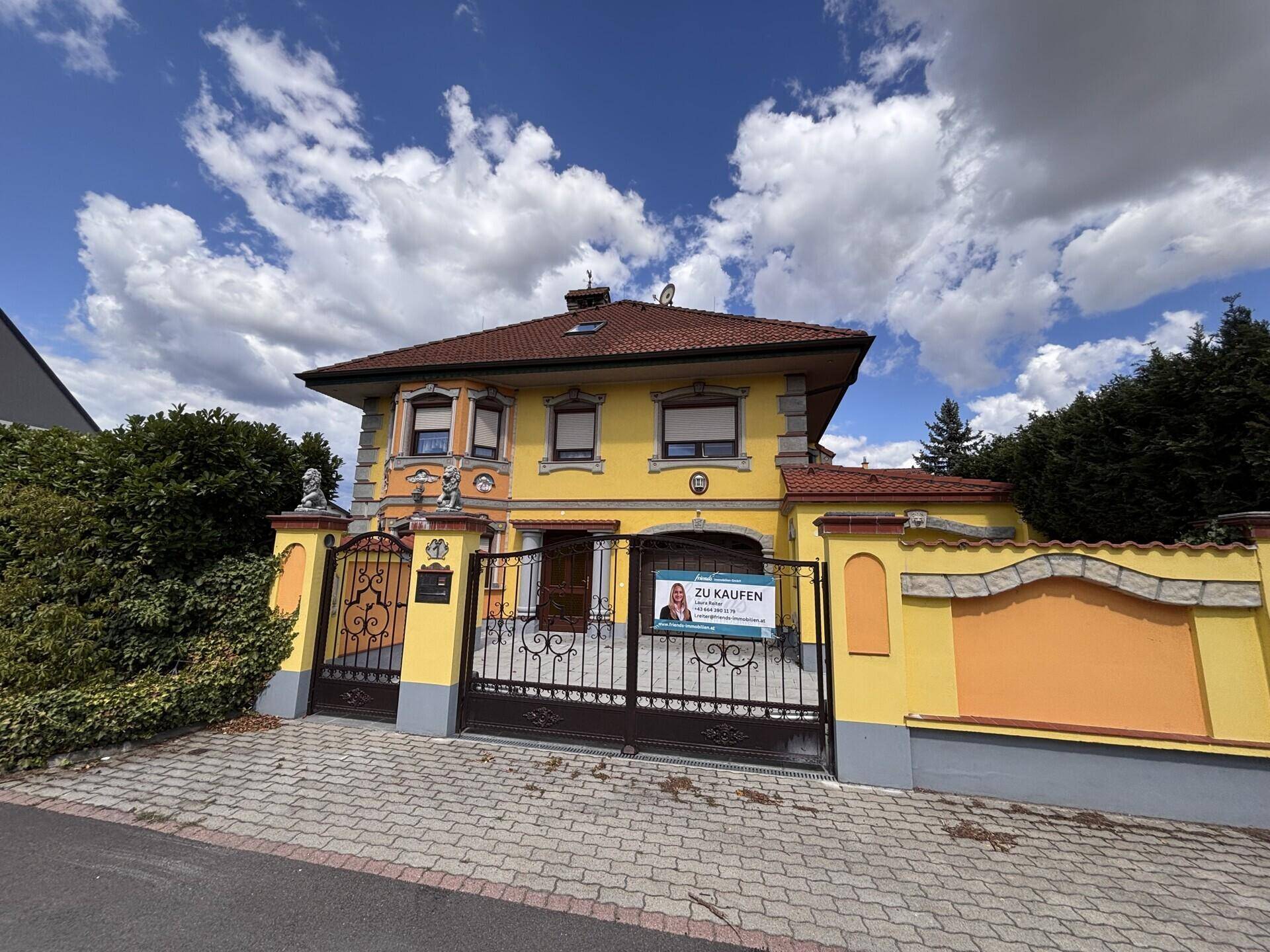 Weitwinkelaufnahme der gelben Hausfassade mit orangefarbenen Details, einem schmiedeeisernen Tor und einem Verkaufsschild.