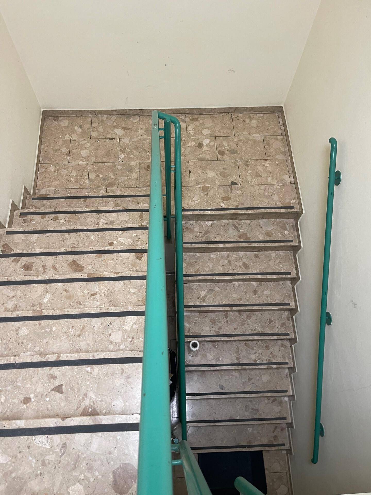 Treppenhaus mit Terrazzoboden und auffälligen grünen Handläufen, die nach unten führen.