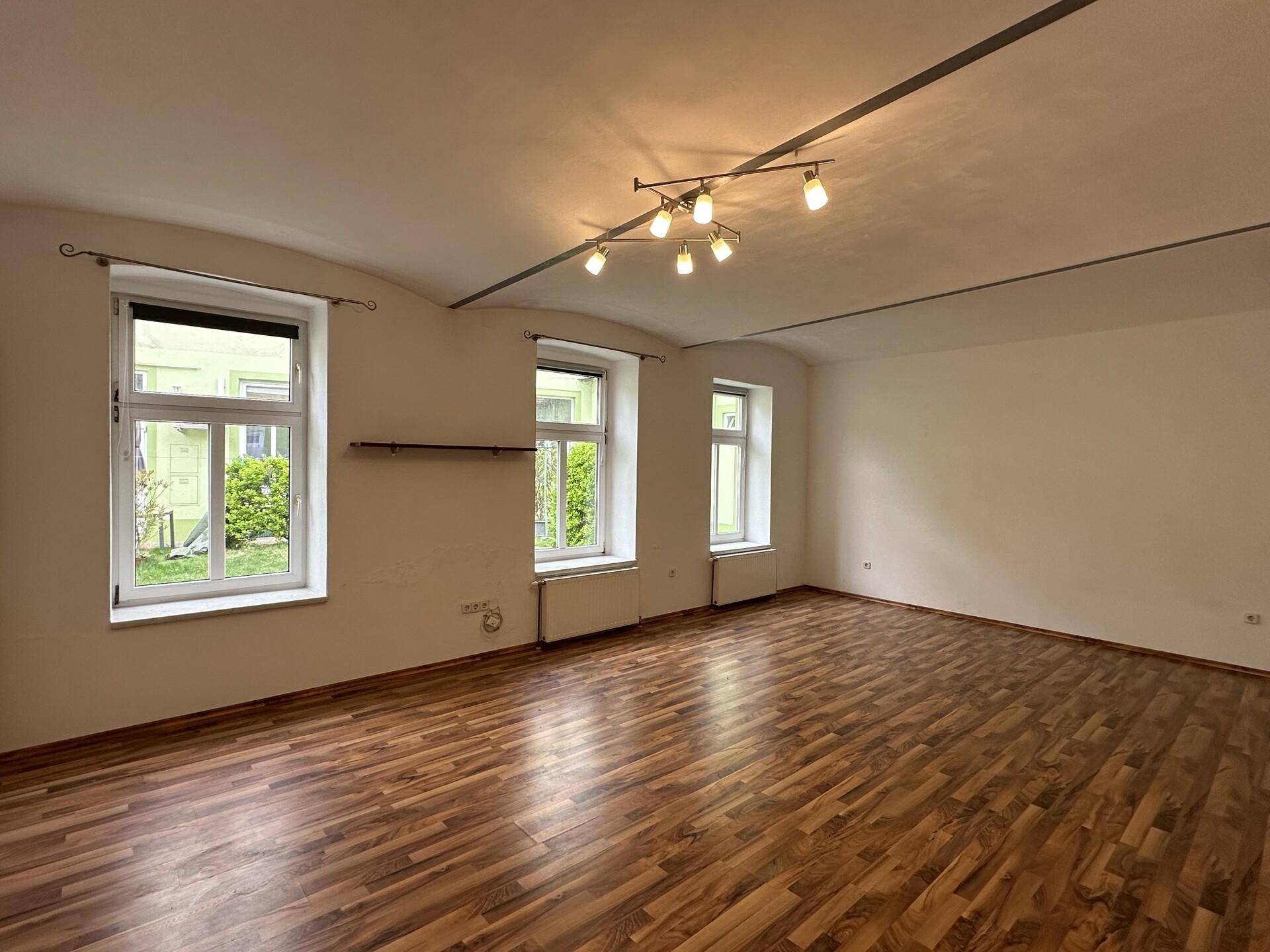 Geräumiges Wohnzimmer mit Holzboden und mehreren Fenstern mit Blick ins Grüne.