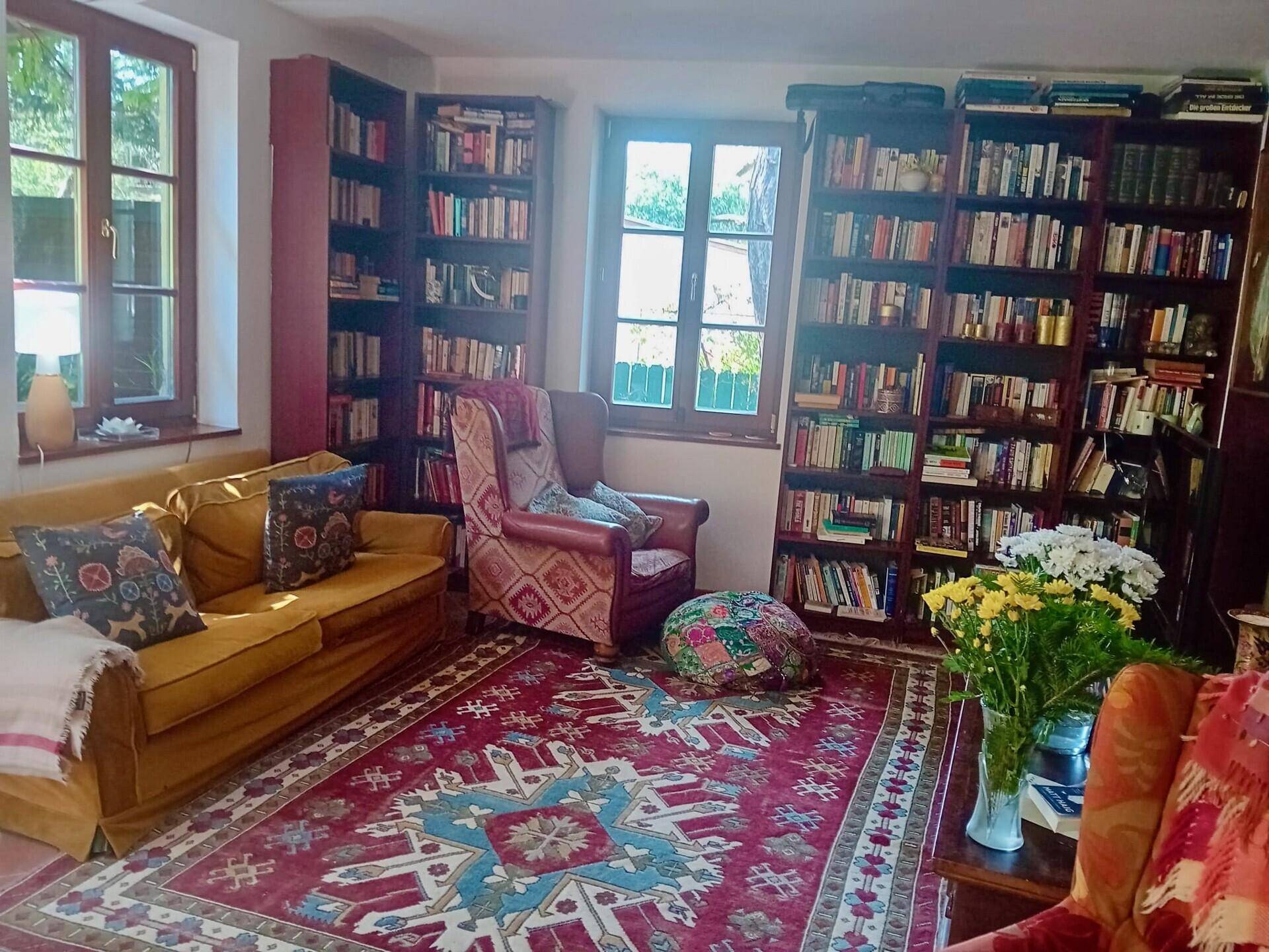 Gemütliches Wohnzimmer mit vielen Bücherregalen, Sofa und Sessel, Teppichboden.