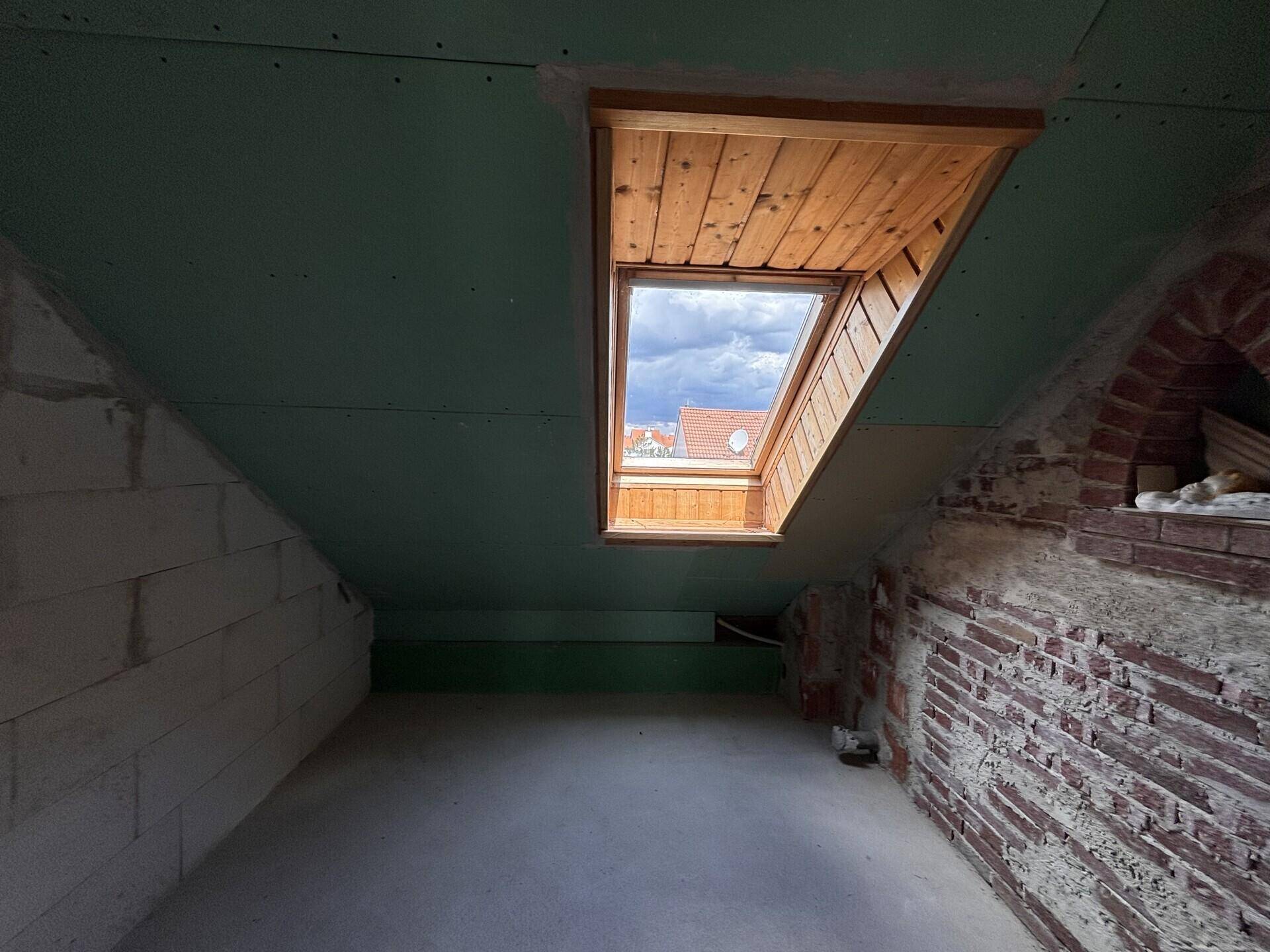 Dachgeschossraum im Rohbau mit Dachfenster und sichtbaren Ziegelwänden.