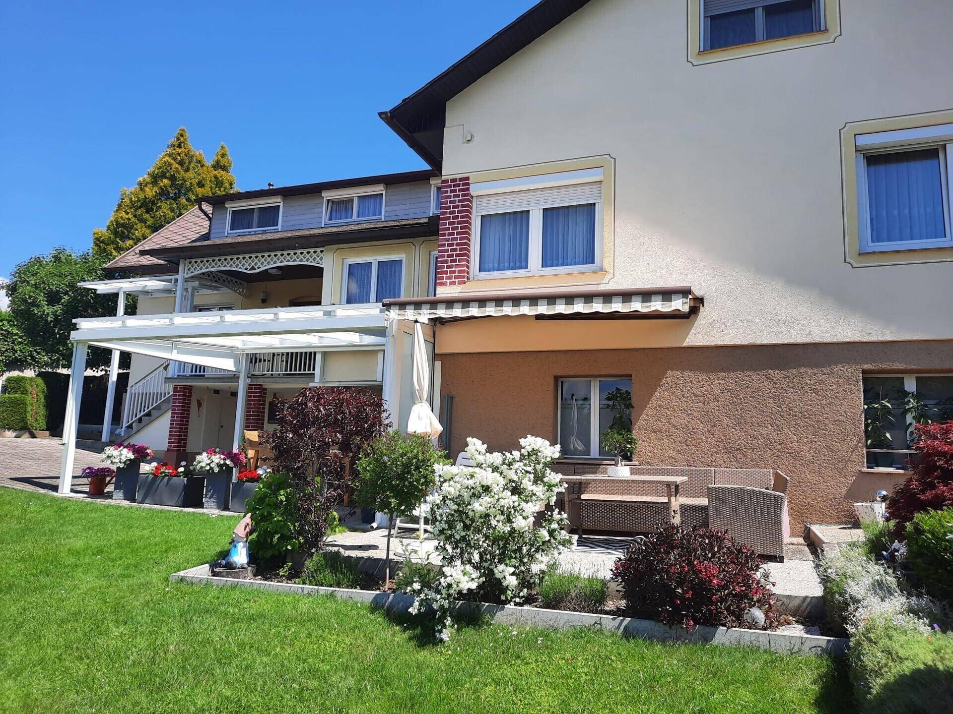 Detailansicht der Hausfassade mit überdachter Terrasse und blühenden Sträuchern im Vorgarten.