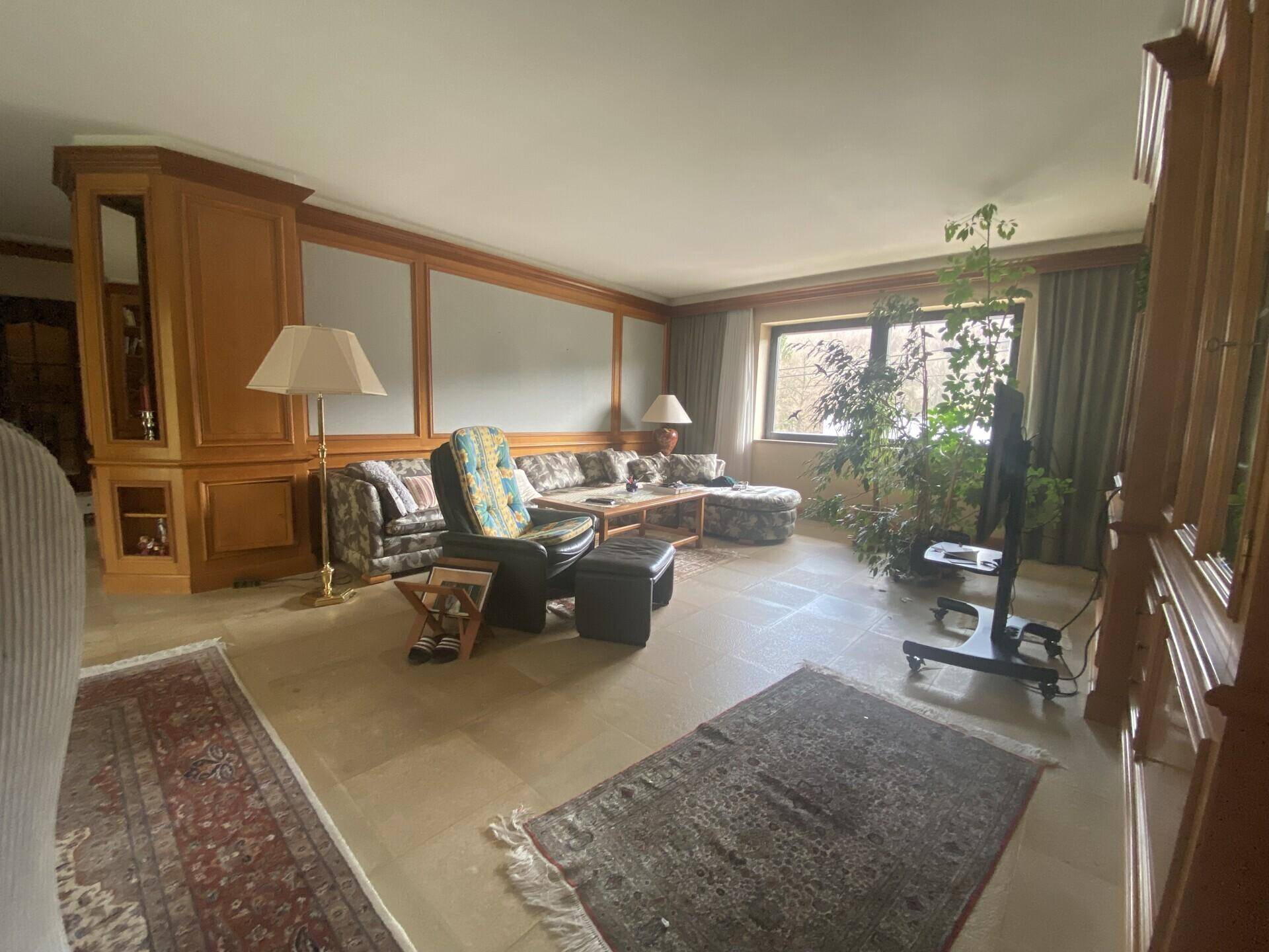 Geräumiges Wohnzimmer mit Fliesenboden, Holzvertäfelung und großen Fenstern mit Blick ins Grüne.
