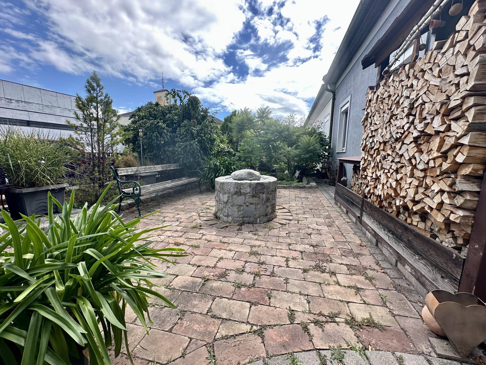 Gepflasterte Terrasse mit Feuerstelle, Holzbank und gestapeltem Brennholz.