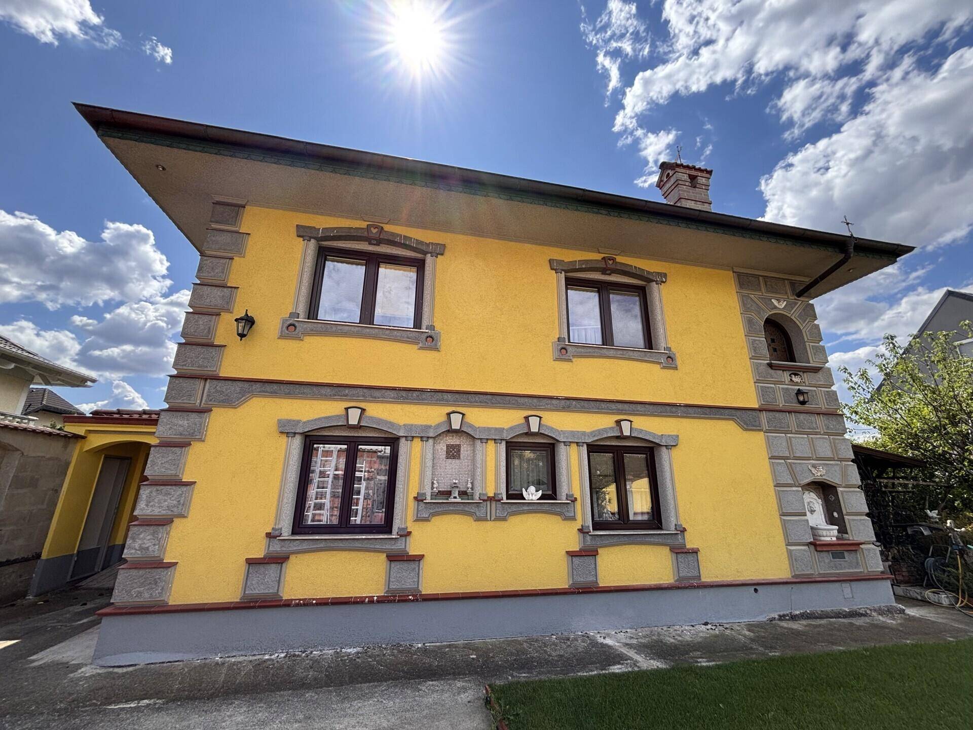 Gelbe Hausfassade mit dekorativen Steinelementen und braunen Fenstern unter blauem Himmel.